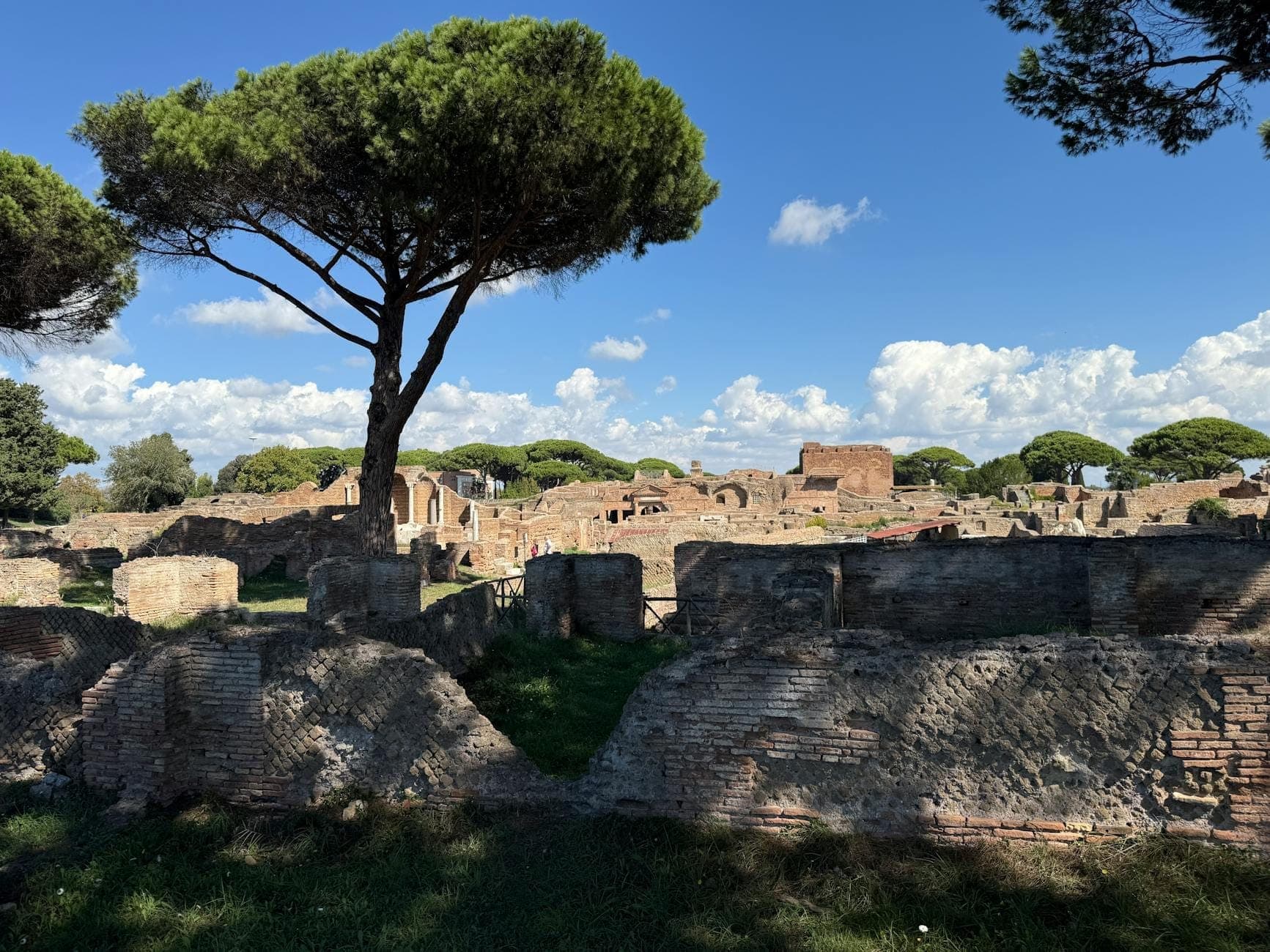 Ruinen und alte Backsteinmauern von Ostia Antica unter blauem Himmel mit Pinien, die das offene und weitläufige Layout der archäologischen Stätte zeigen.