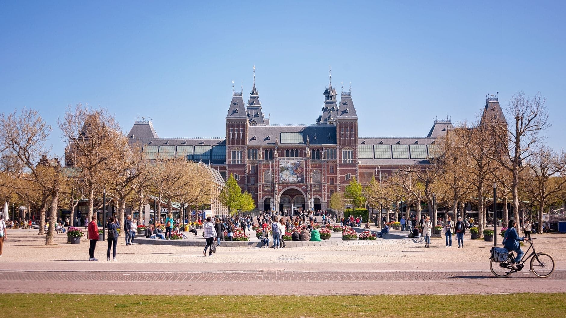Das Rijksmuseum im Stadtteil Oud-Zuid, Amsterdam, steht hinter blattlosen Bäumen und einem belebten Platz voller Menschen an einem sonnigen Tag.