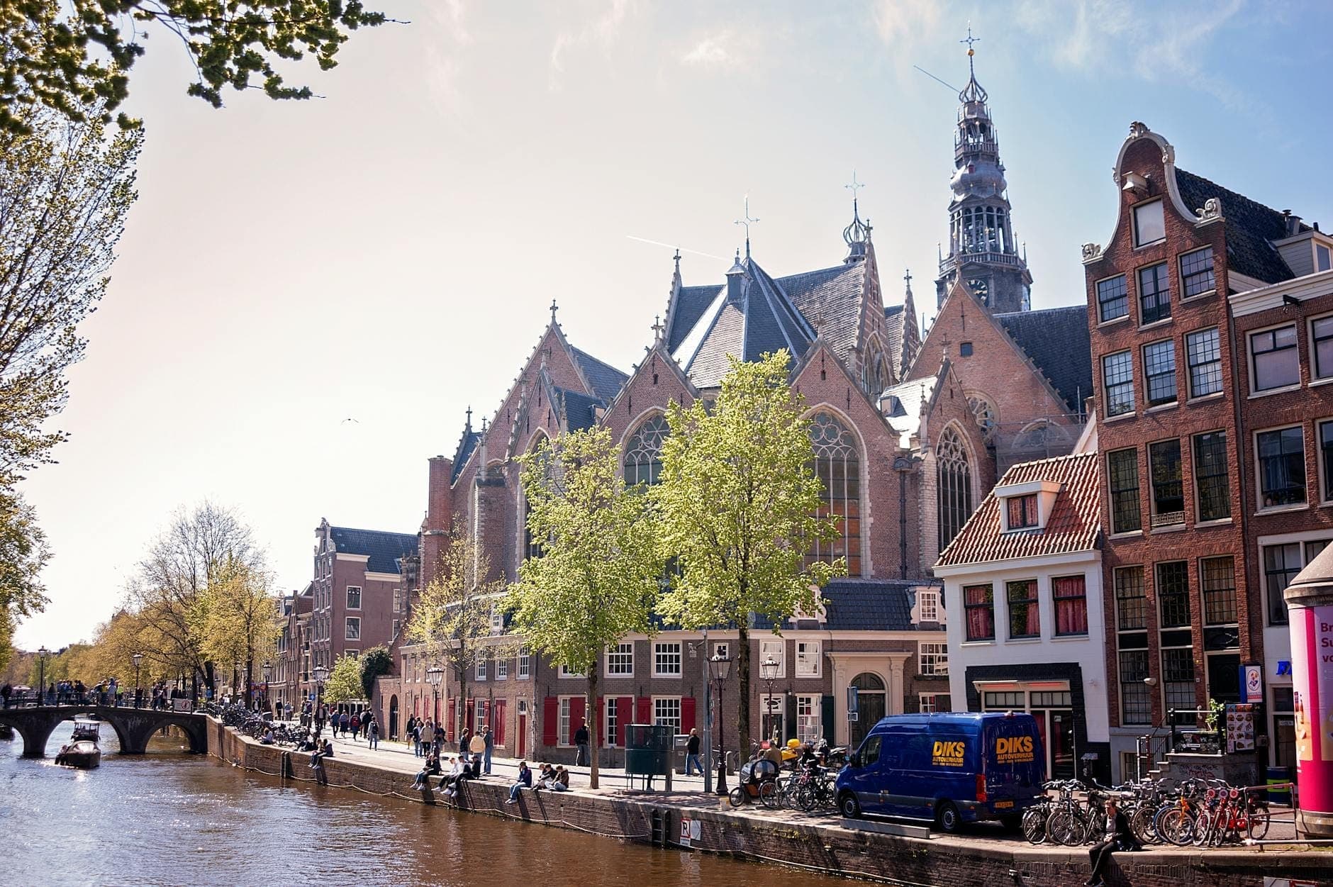 Blick auf die gotische Architektur der Oude Kerk an einem Amsterdamer Kanal, umgeben von historischen Stadtgebäuden und baumgesäumten Straßen unter hellem Himmel.
