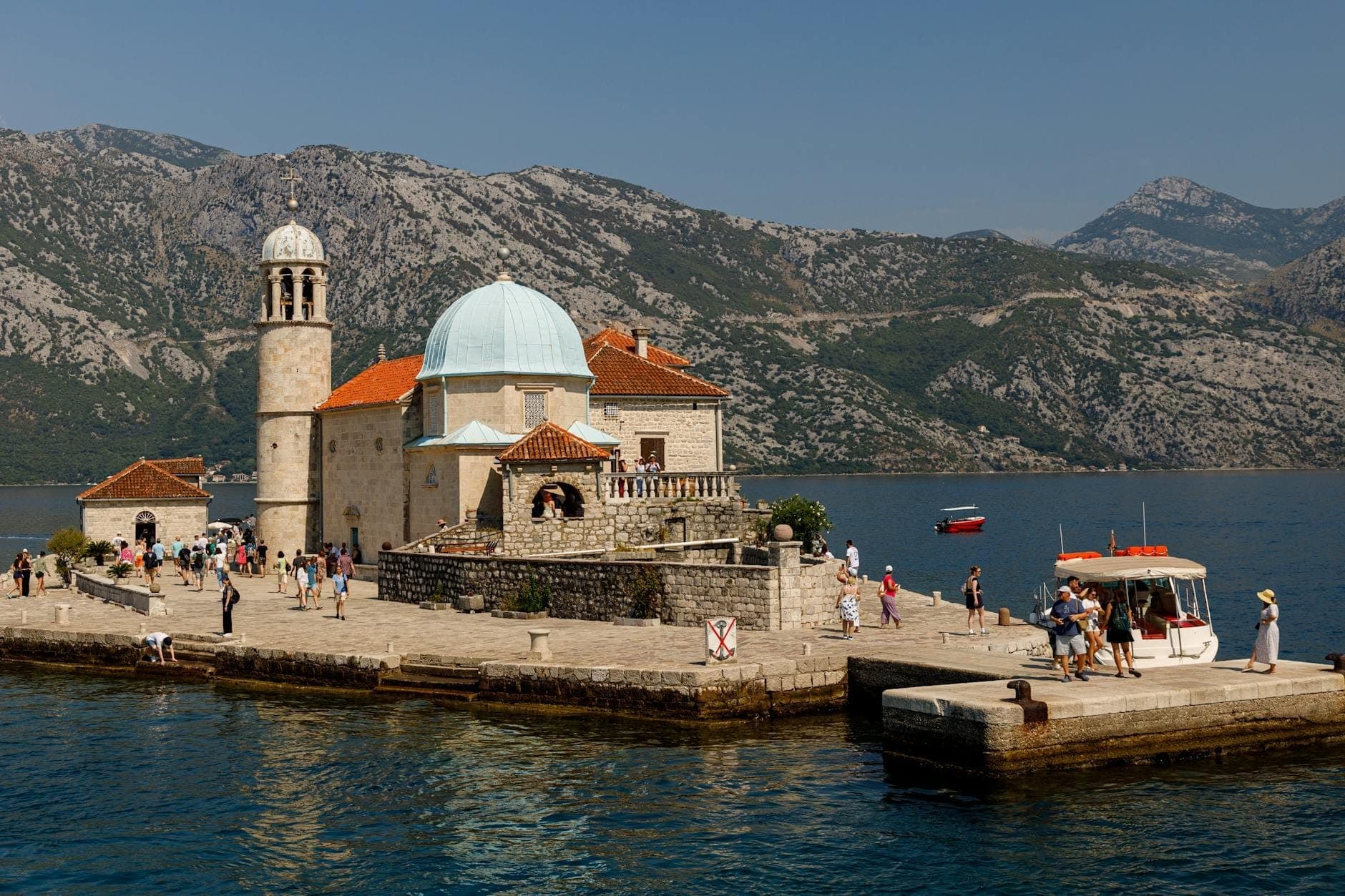 L'église Notre-Dame-du-Rocher avec son dôme bleu et son clocher en pierre sur une île artificielle, entourée de touristes, de bateaux et des montagnes spectaculaires de la baie de Kotor.
