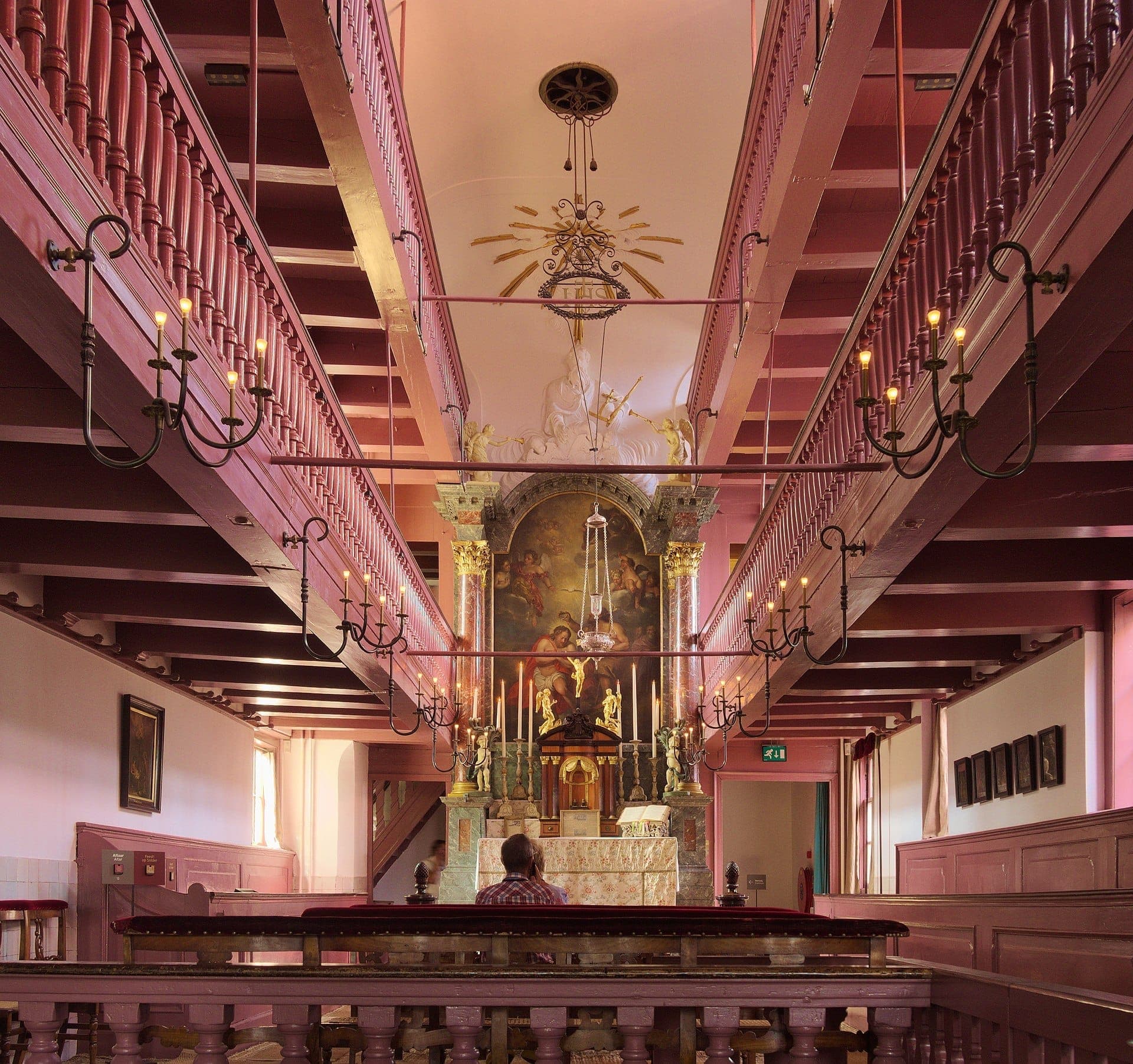 Innenansicht der Kirche des Museums Ons' Lieve Heer op Solder mit verziertem Altar, rosafarbenen Holzbalkonen, Kronleuchtern und einem sitzenden Besucher.