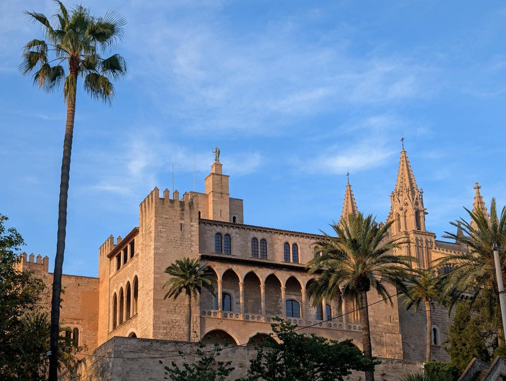 Der Palast La Almudaina in Palma, Mallorca, ist mit seinen Steinmauern, Zinnen und Palmen unter einem klaren blauen Himmel zu sehen.