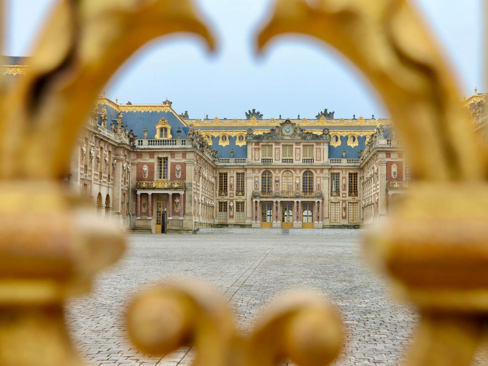 The main entrance to the Palace of Versailles framed by ornate golden gate decoration, with a wide cobblestone courtyard leading to the iconic palace façade.
