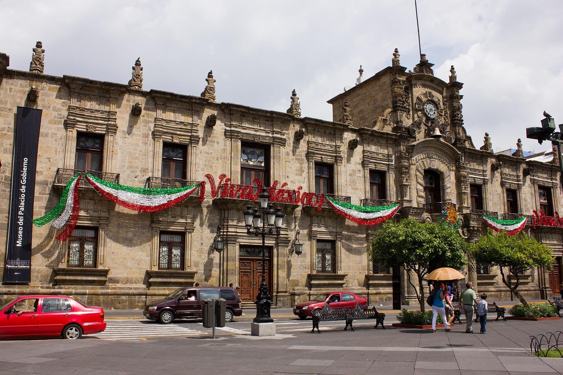 ด้านนอกของ Palacio de Gobierno de Jalisco ที่มีประวัติศาสตร์ ประดับด้วยธงชาติเม็กซิโกเทศกาลและป้าย 'Viva México' มีรถยนต์และคนเดินเท้าอยู่ด้านหน้า