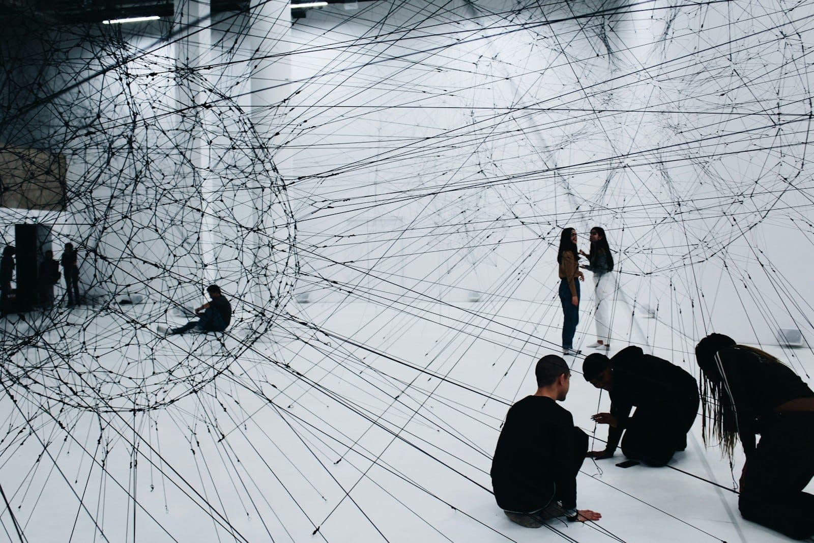 People interacting with a large-scale, immersive contemporary art installation made of black string in a white exhibition space.