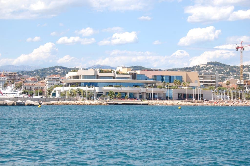 Здание Palais des Festivals et des Congrès, вид с воды, с пальмами, солнечным небом и панорамой Канн на заднем плане.