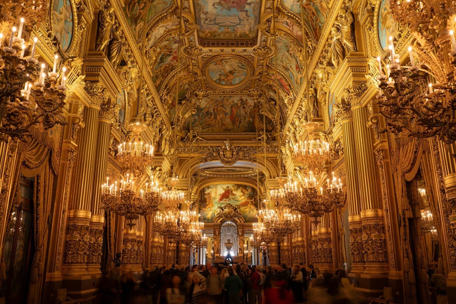 Lavishly decorated grand hall inside the Palais Garnier with ornate gold details, chandeliers, and a painted ceiling, full of visitors admiring the architecture.