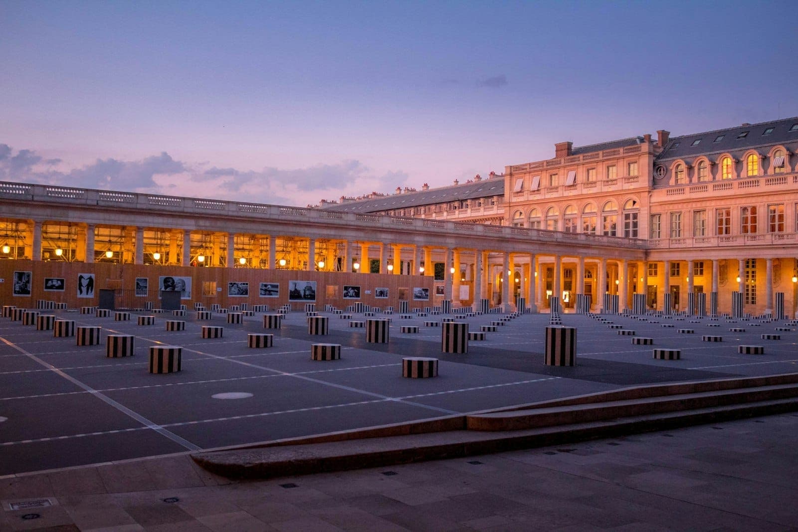 The Palais Royal courtyard at dusk with the iconic black and white Buren columns, classical arcades, and warm building lights under a purple sky.