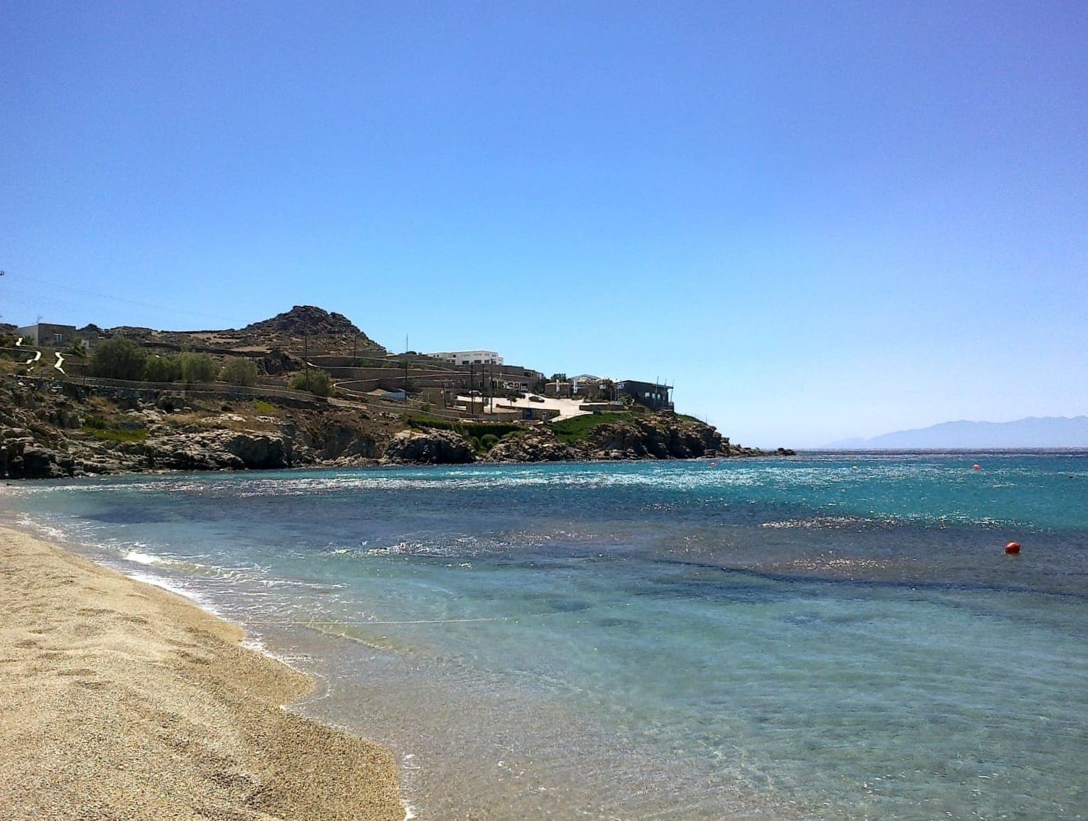 Klares blaues Wasser und goldener Sand säumen den Paradise Beach, Mykonos, mit felsigen Hügeln und weißen Gebäuden unter einem sonnigen Himmel.