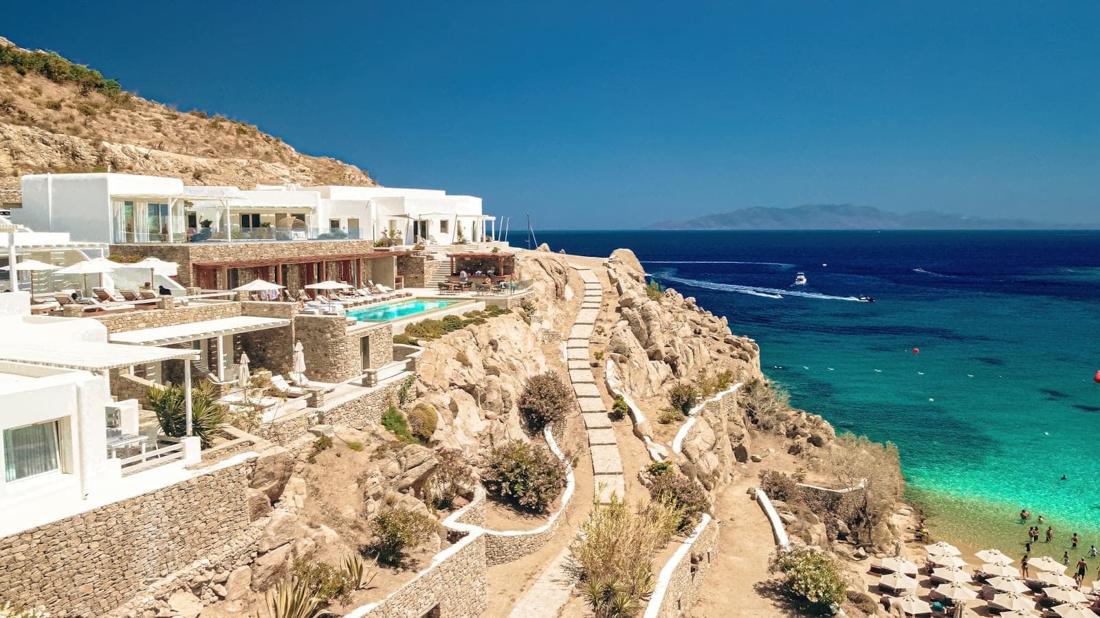 Blick von der Klippe auf Paradise oder Super Paradise Beach auf Mykonos mit türkisfarbenem Wasser, Sonnenliegen, weißen Luxusgebäuden und strahlend blauem Himmel.