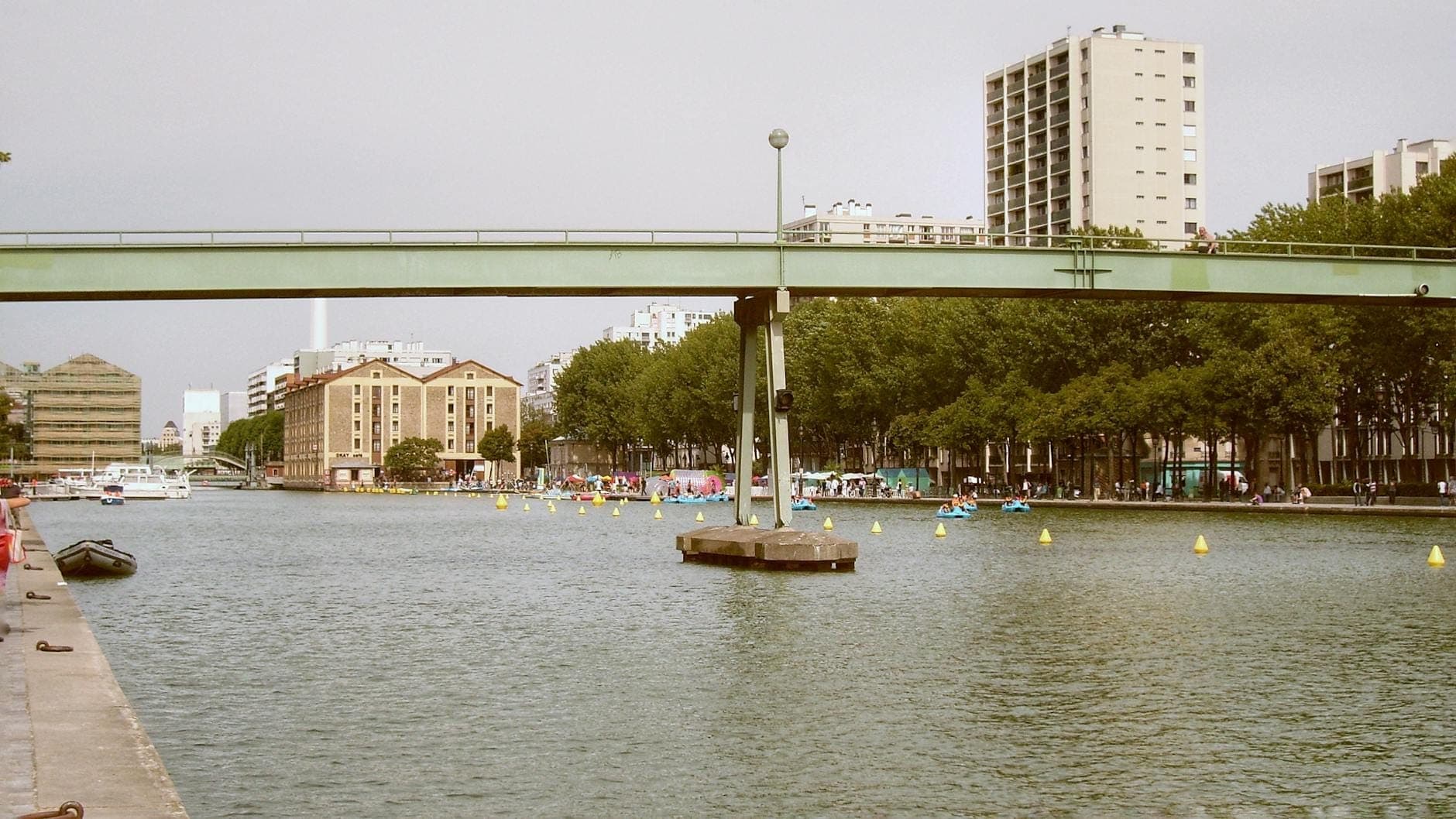 A wide canal in an urban park with a footbridge, tree-lined paths, modern buildings, and people enjoying the park on both sides of the water.