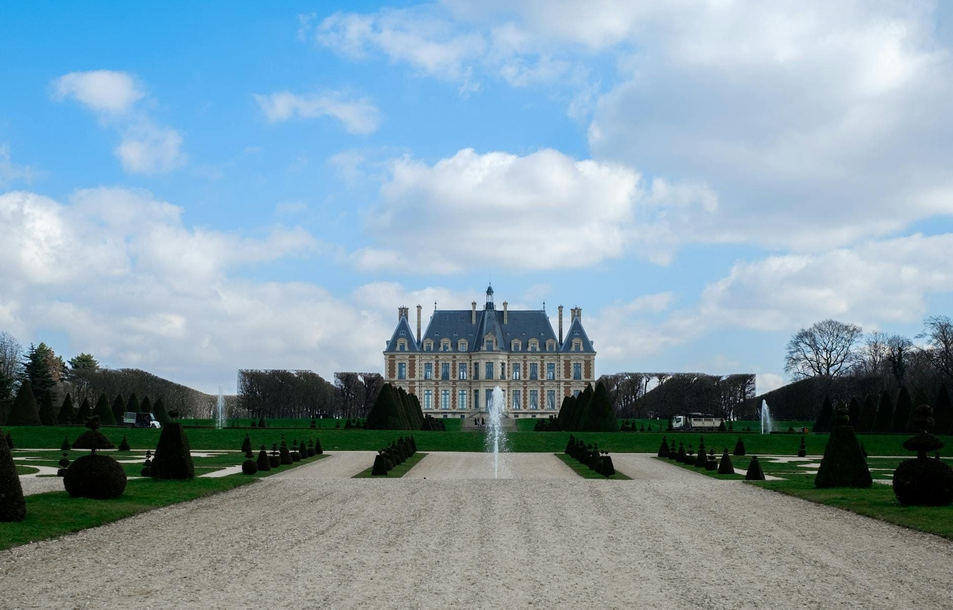 Wide view of the château at Parc de Sceaux framed by formal French gardens, symmetrical pathways, and a central fountain under a blue sky with clouds.
