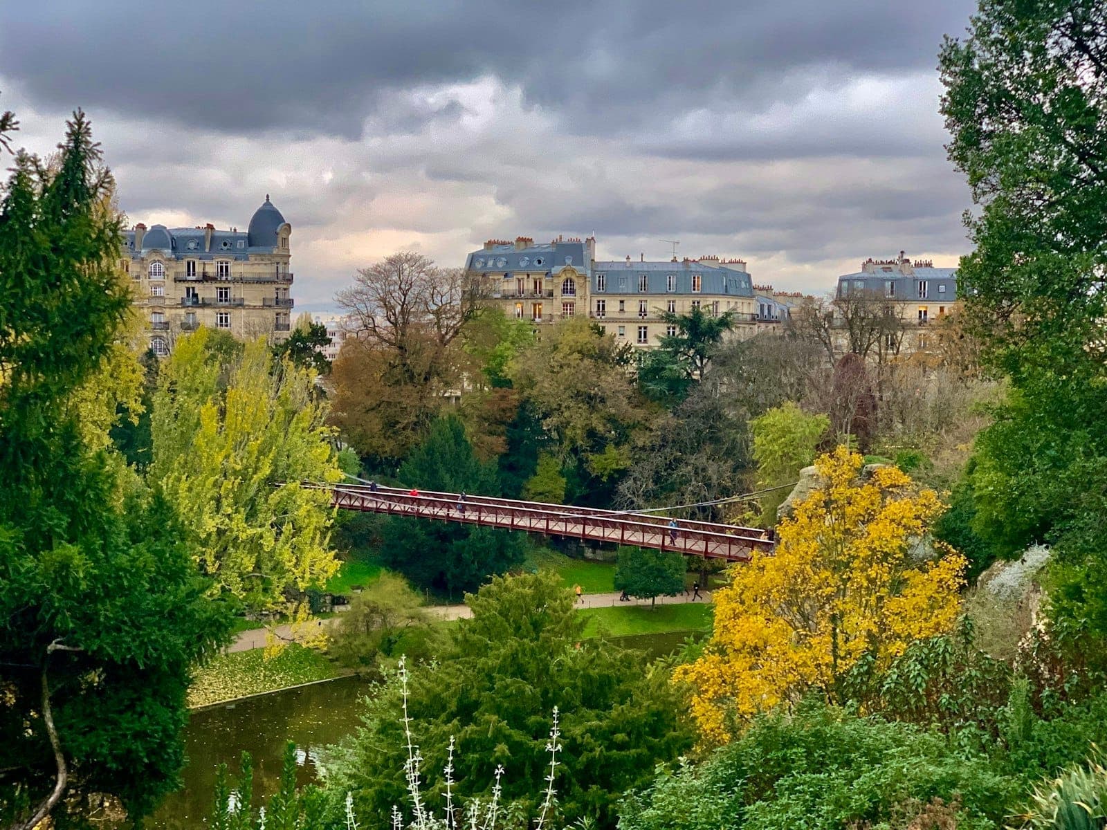 Dramatic view of Parc des Buttes-Chaumont with lush green trees, a red suspension bridge, and classic Parisian buildings under a cloudy sky.