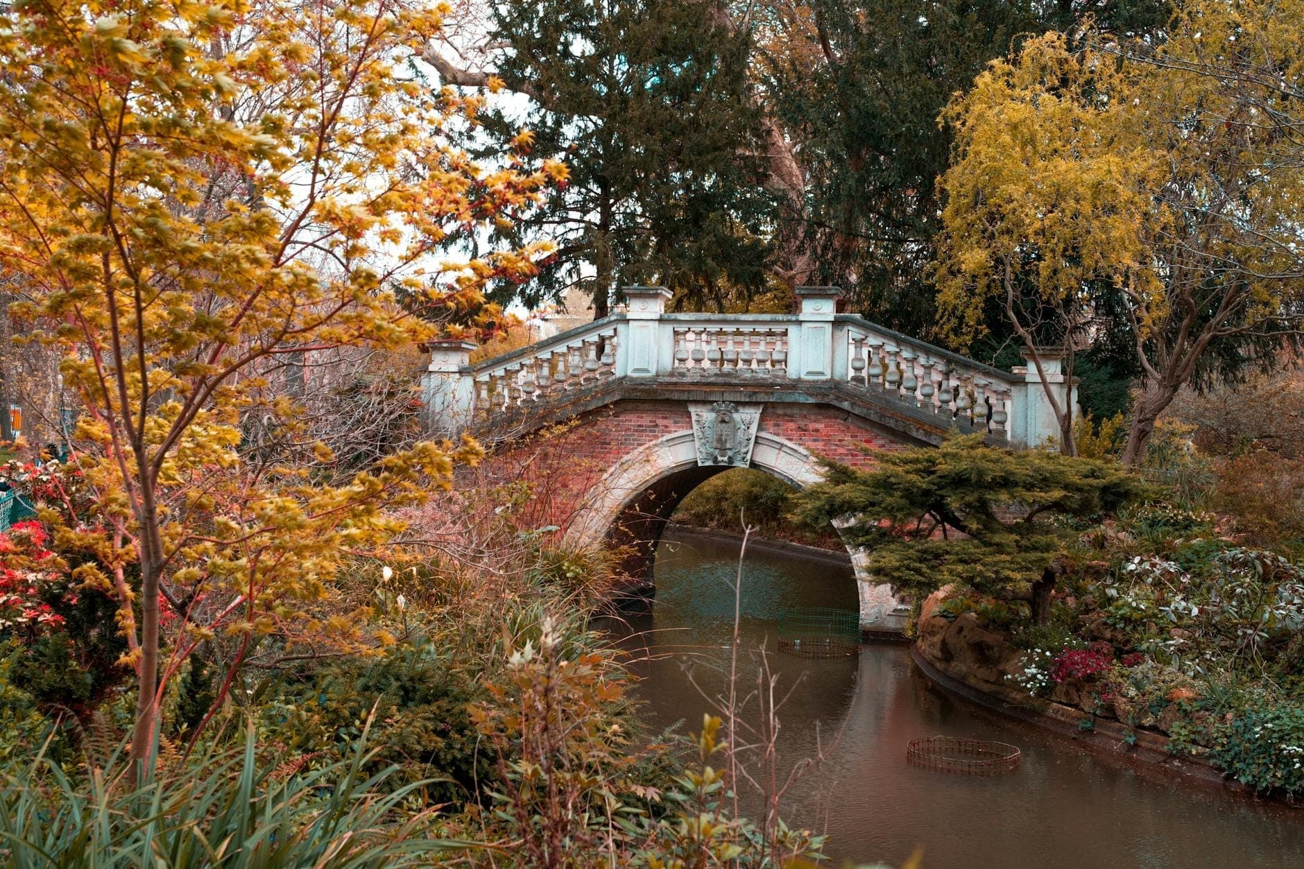Picturesque arched stone bridge over a tranquil artificial pond surrounded by lush trees and colorful autumn foliage in a peaceful city park setting.