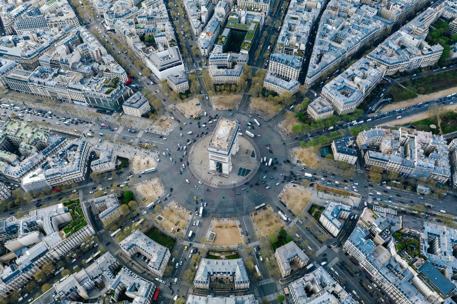 Aerial view of the Arc de Triomphe at the center of a star-shaped road layout surrounded by classic Parisian buildings.