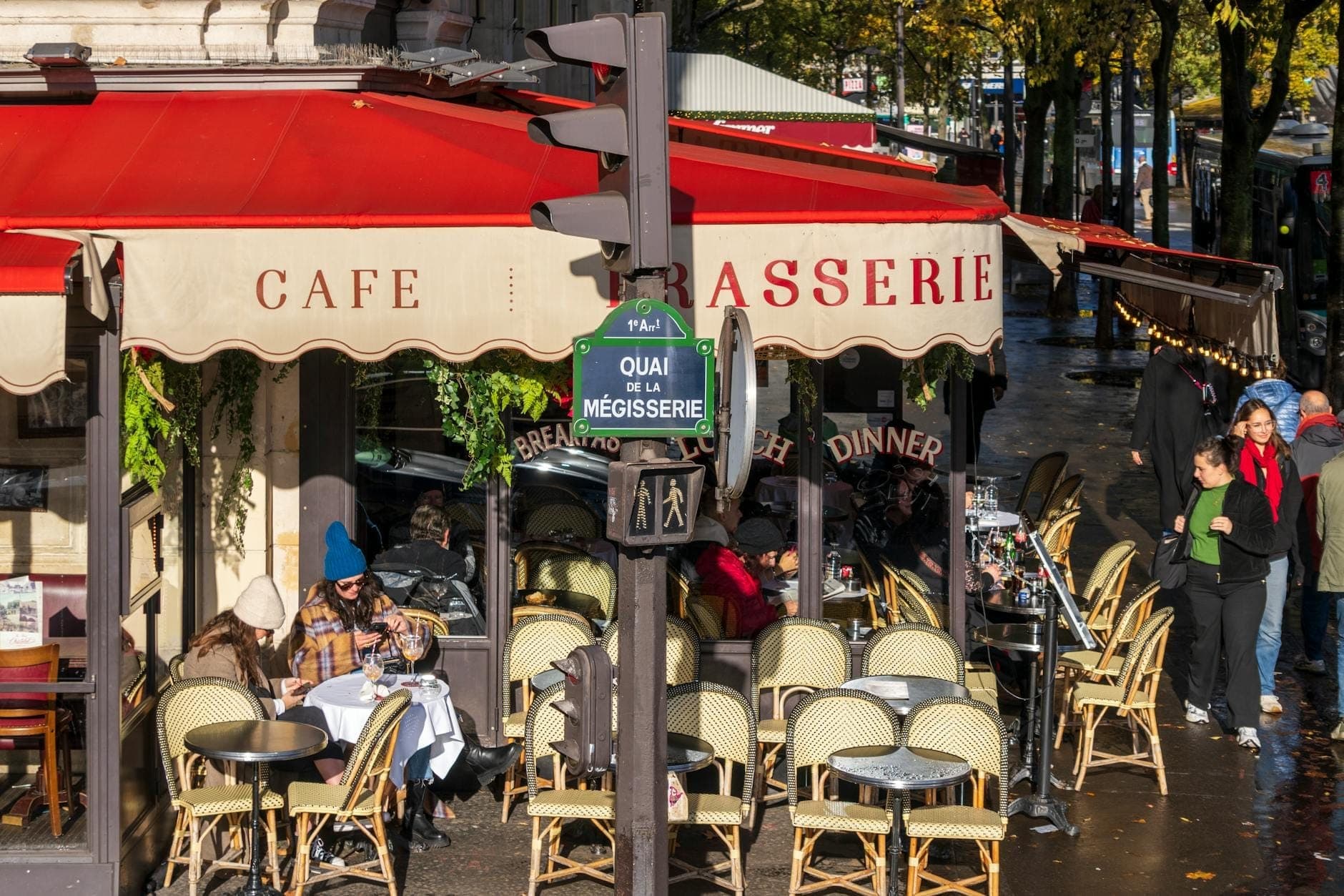 Quán cà phê vỉa hè Paris cổ điển với mái hiên màu đỏ, chỗ ngồi ngoài trời và mọi người dùng bữa tại các bàn bên cạnh vỉa hè có hàng cây xanh.