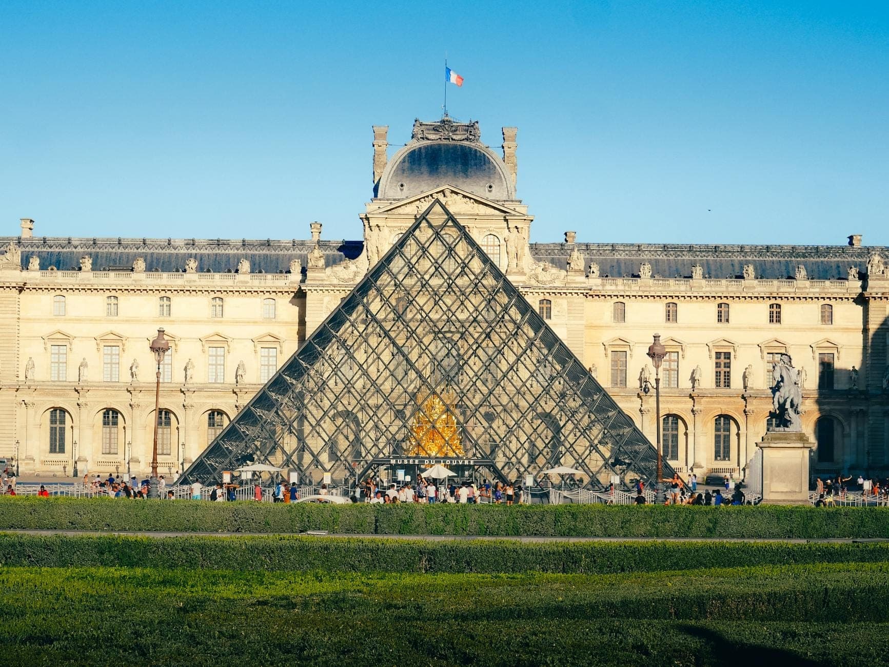 Cảnh toàn cảnh kim tự tháp kính của Louvre và lối vào bảo tàng với mọi người xếp hàng bên ngoài dưới bầu trời xanh trong.
