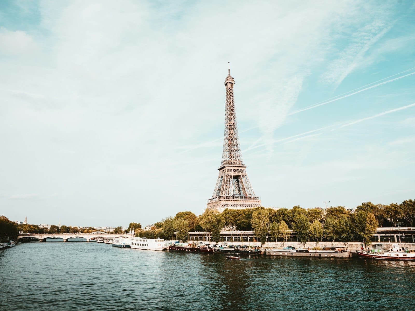 Wide view of the Eiffel Tower beside the Seine River on a clear day, with boats, trees, and Paris cityscape in the background.