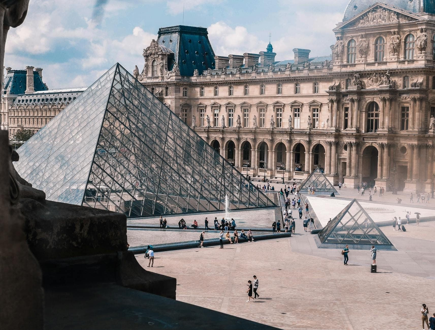 Louvre Müzesi avlusunun, ikonik cam piramidi ve çevresindeki tarihi binalarla birlikte, meydanda yürüyen ziyaretçileri gösteren fotoğrafı.