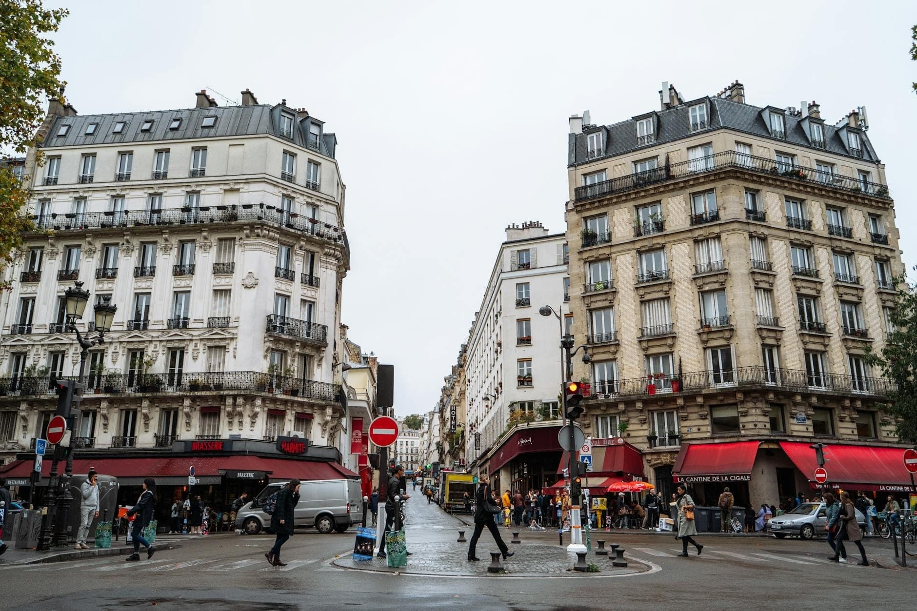 A lively Parisian street scene with classic buildings, people walking, and bustling cafes with red awnings on a cloudy day.