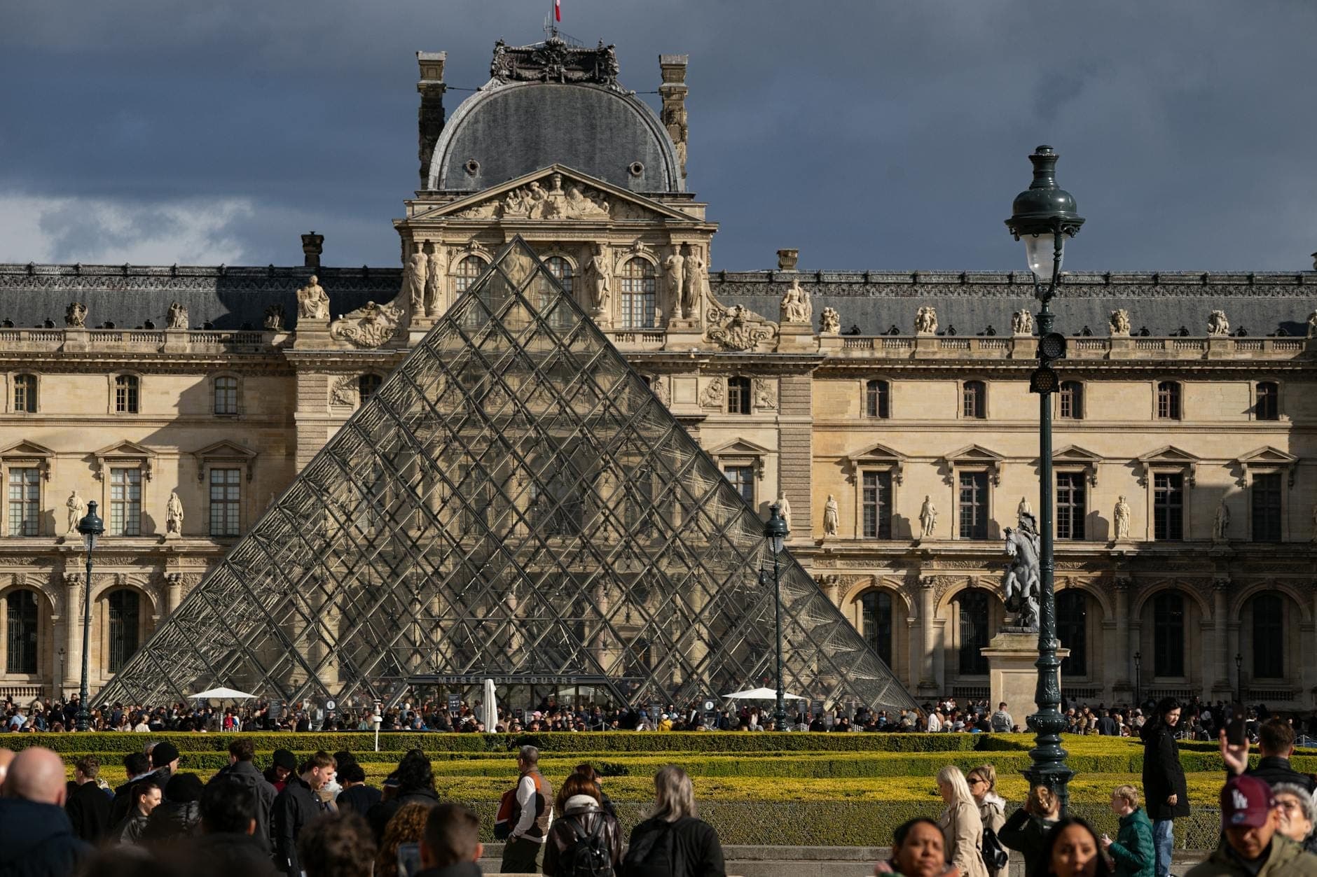 พิพิธภัณฑ์ลูฟวร์และทางเข้าพีระมิดกระจกอันเป็นสัญลักษณ์ในปารีส พร้อมนักท่องเที่ยวจำนวนมากในเบื้องหน้าภายใต้ท้องฟ้าที่งดงาม