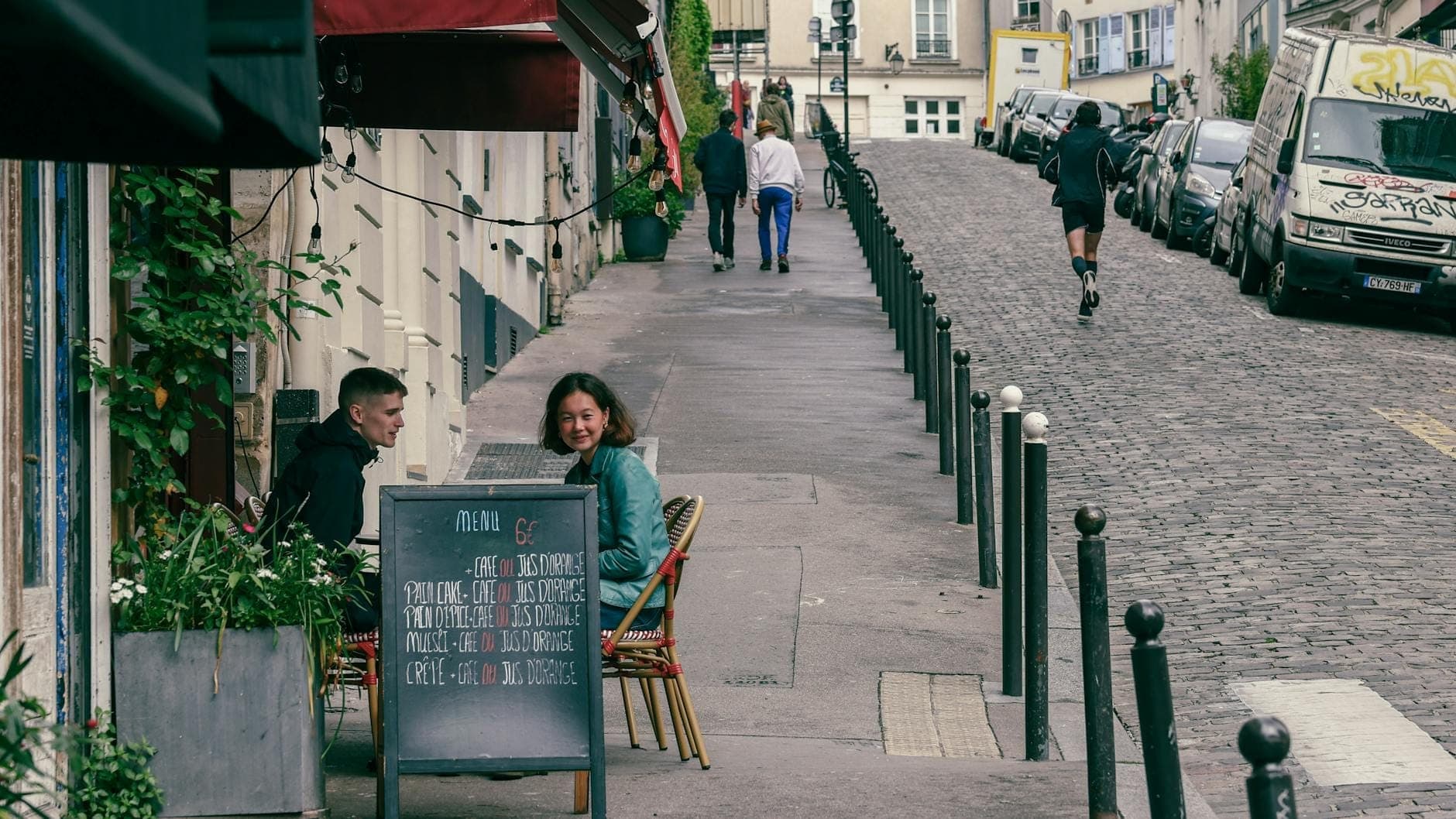 ภาพถนนในปารีสพร้อมผู้คนที่นั่งรับประทานอาหารที่คาเฟ่กลางแจ้งเล็กๆ กระดานเมนูพร้อมราคาด้านหน้า และสถาปัตยกรรมแบบฉบับของปารีสเป็นฉากหลัง