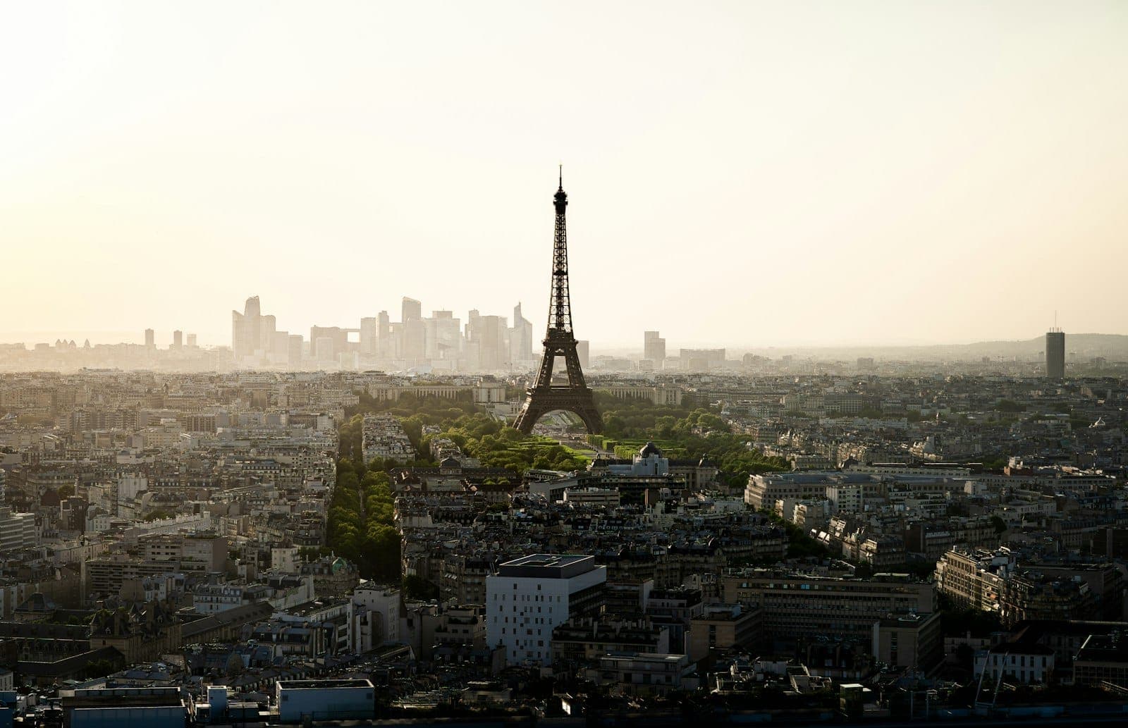A sweeping view of Paris at sunset with the Eiffel Tower prominently in the center, framed by city buildings and a hazy skyline.