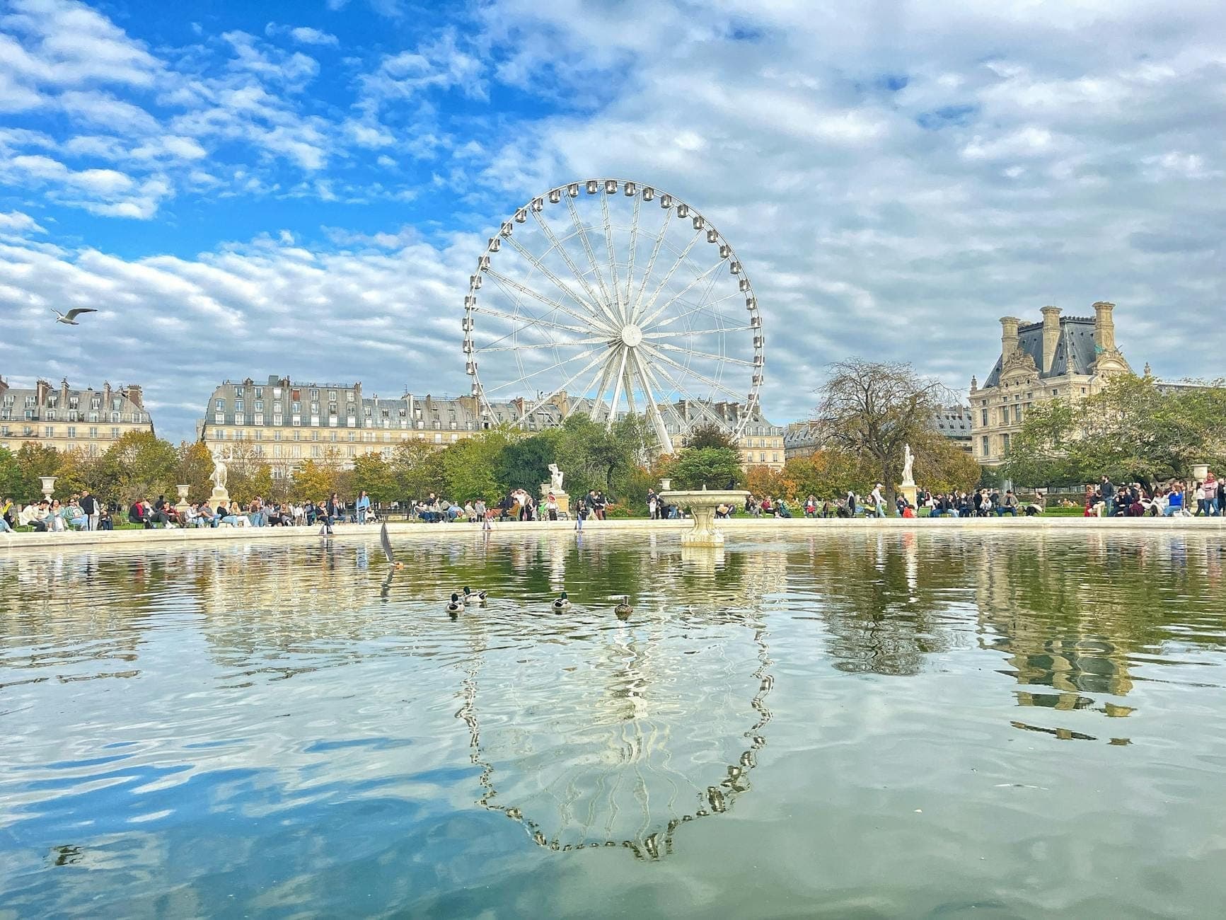 Helle, lebhafte Szene im Jardin des Tuileries in Paris mit einem großen Riesenrad, einem Teich, klassischen Gebäuden sowie Kindern und Familien, die die Natur genießen.