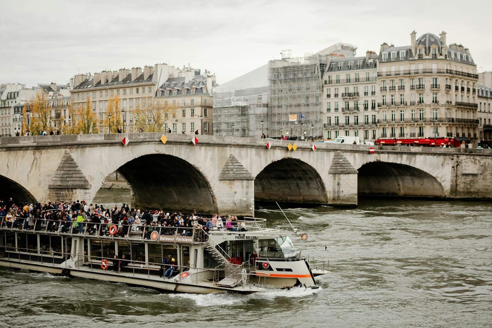 Ein langer Ausflugsboot voller Menschen fährt unter einer historischen Steinbrücke auf der Seine in Paris hindurch, mit Pariser Gebäuden im Hintergrund.