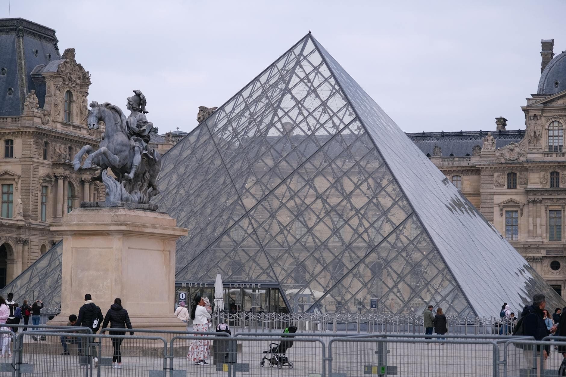 Blick auf den Eingang der Louvre-Pyramide in Paris mit Familien, einer Statue im Vordergrund und Menschen, die über den Platz spazieren.