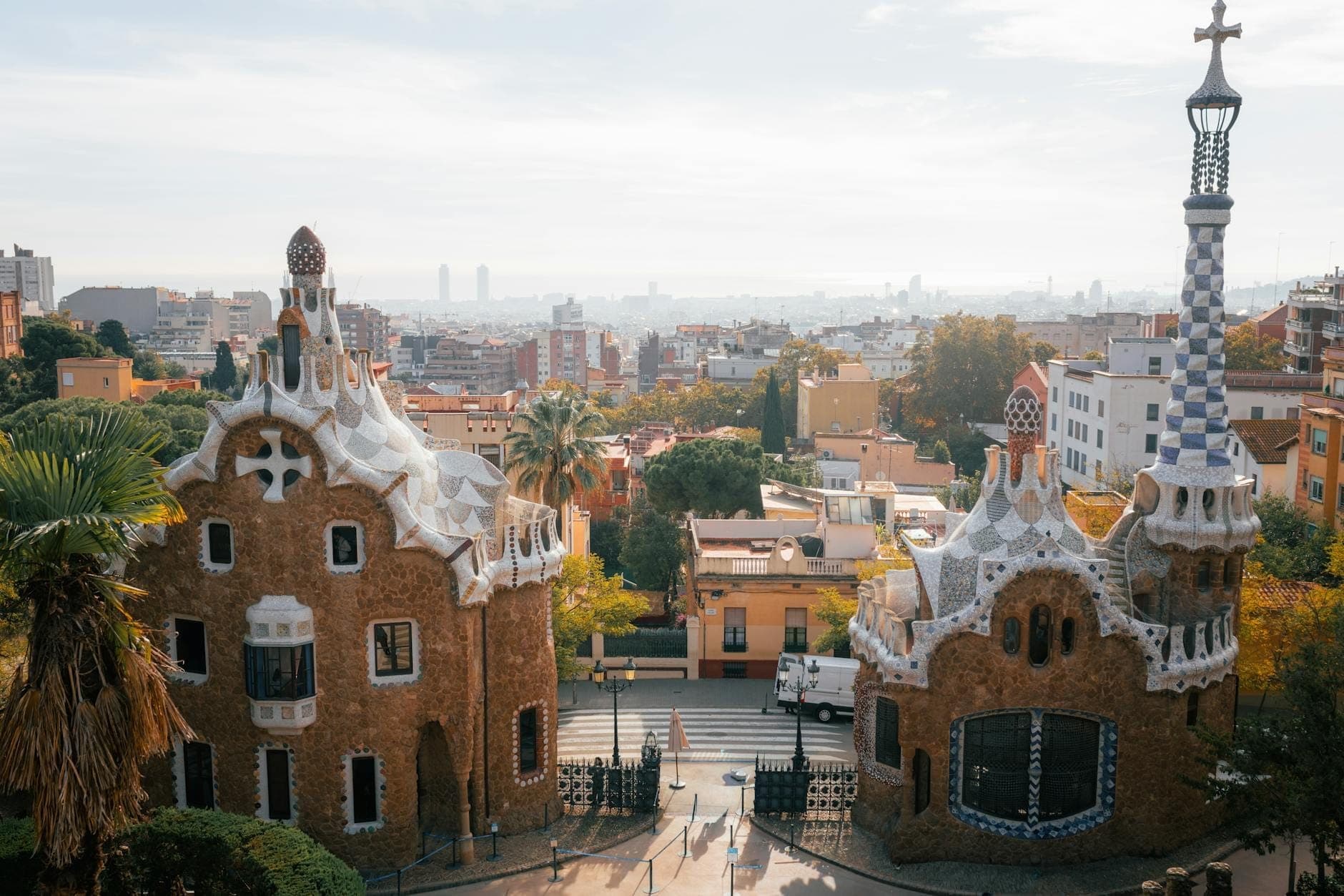 Vista panorâmica ampla da entrada do Park Güell, com as encantadoras casas de portão de Gaudí e o horizonte de Barcelona se estendendo até o Mediterrâneo em um dia de sol.