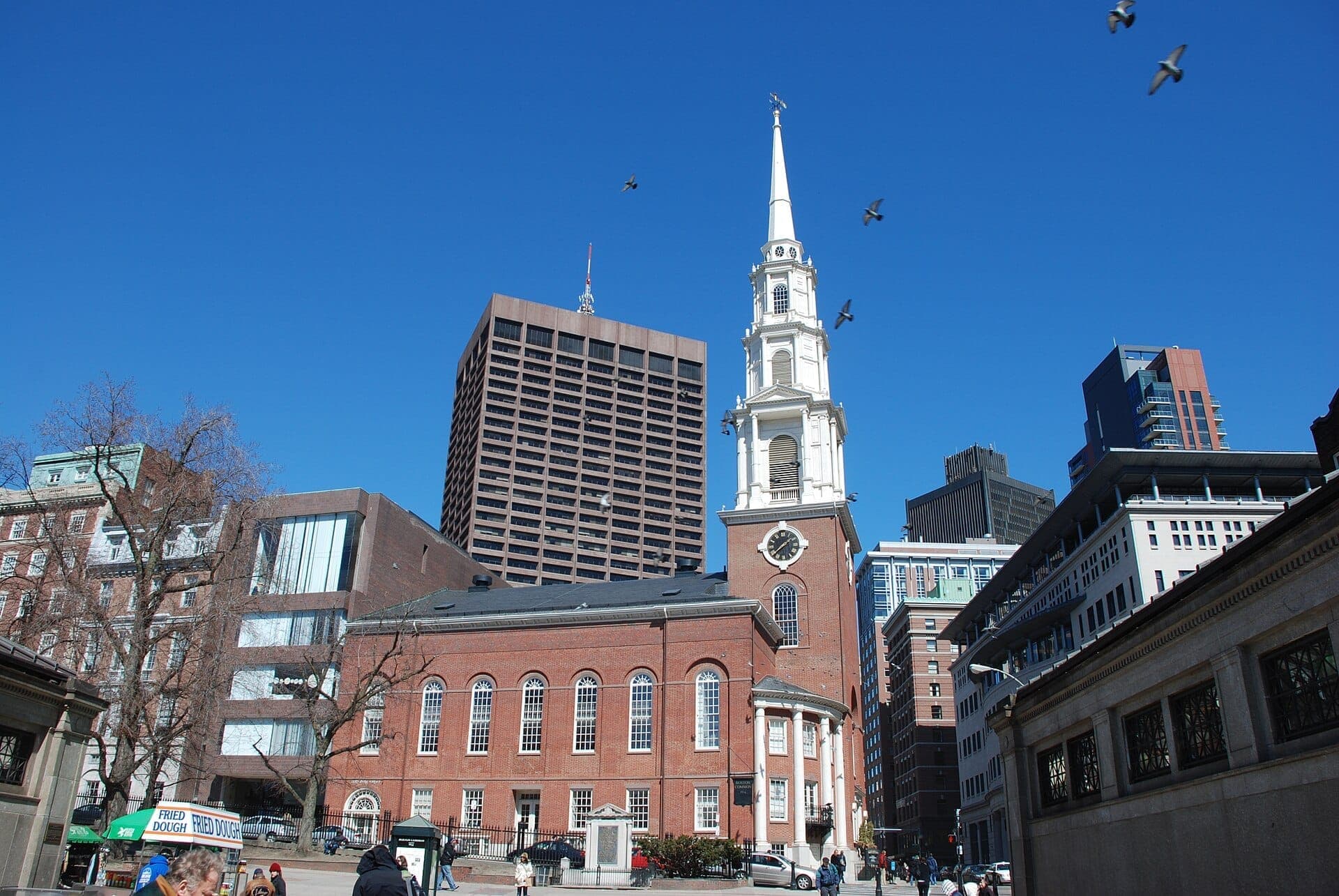 La Park Street Church avec son clocher blanc se distingue parmi les immeubles modernes de Boston sous un ciel bleu dégagé, avec des passants à proximité.