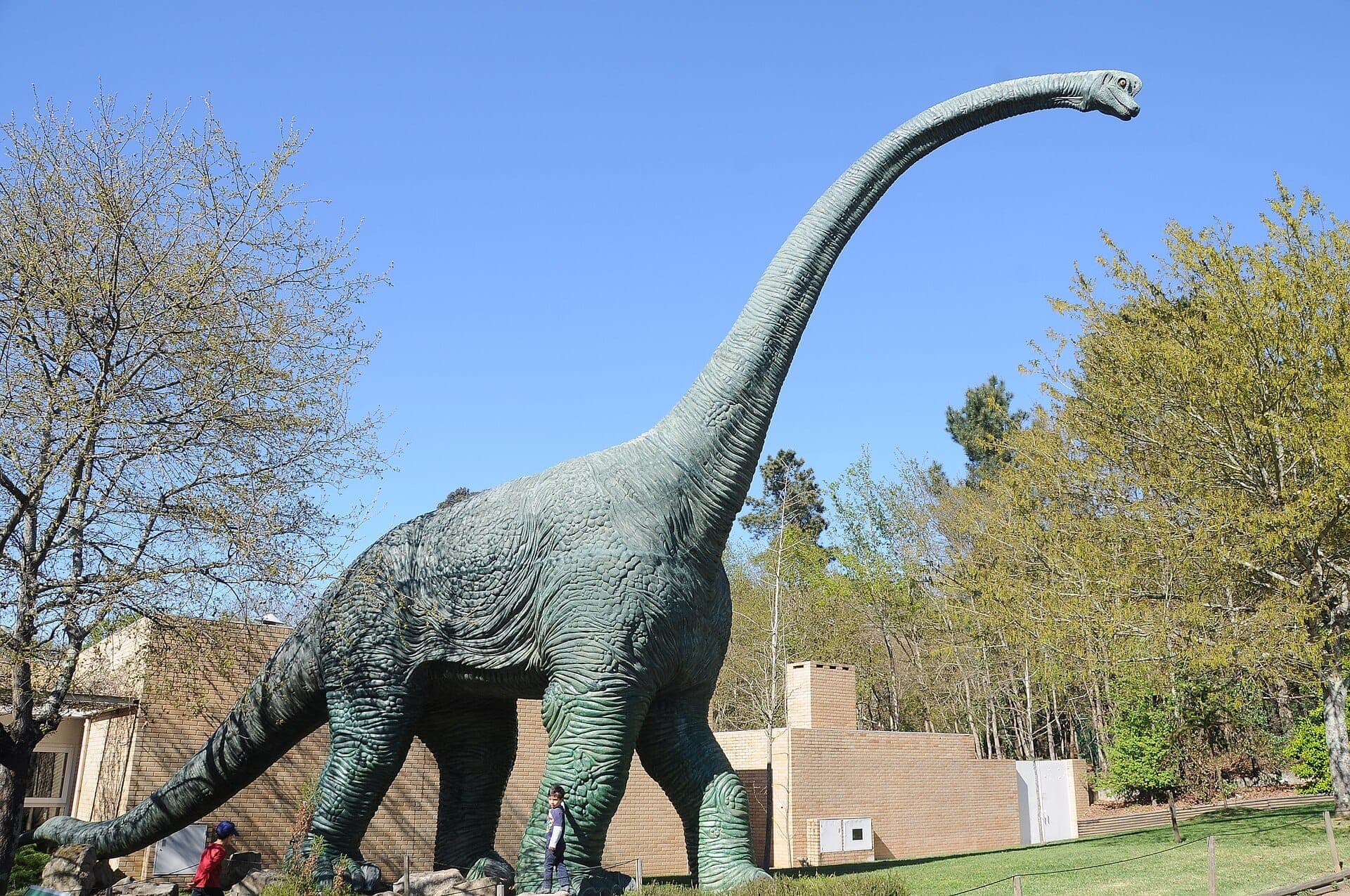 Duży posąg dinozaura w Parque Biológico de Gaia otoczony wiosennymi drzewami i ceglanymi budynkami pod bezchmurnym błękitnym niebem.