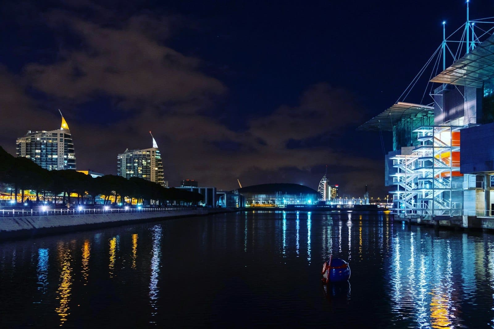 Vista noturna da frente ribeirinha do Parque das Nações com prédios modernos, torres icônicas e o Oceanário de Lisboa refletido no rio sob um céu dramático.