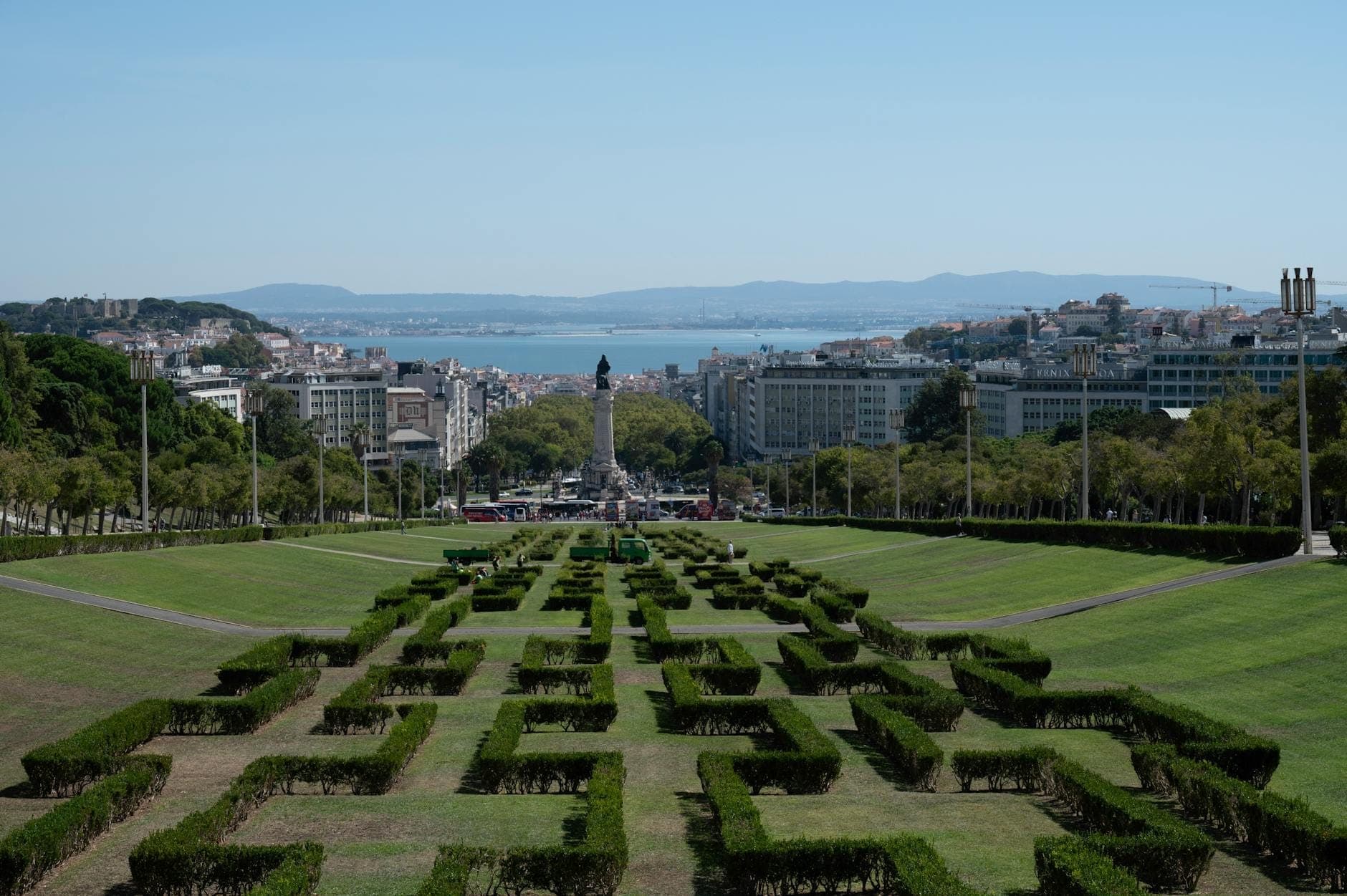 Vista das sebes geométricas do Parque Eduardo VII, a avenida central e o horizonte de Lisboa com o Rio Tejo e as colinas ao fundo sob um céu azul límpido.