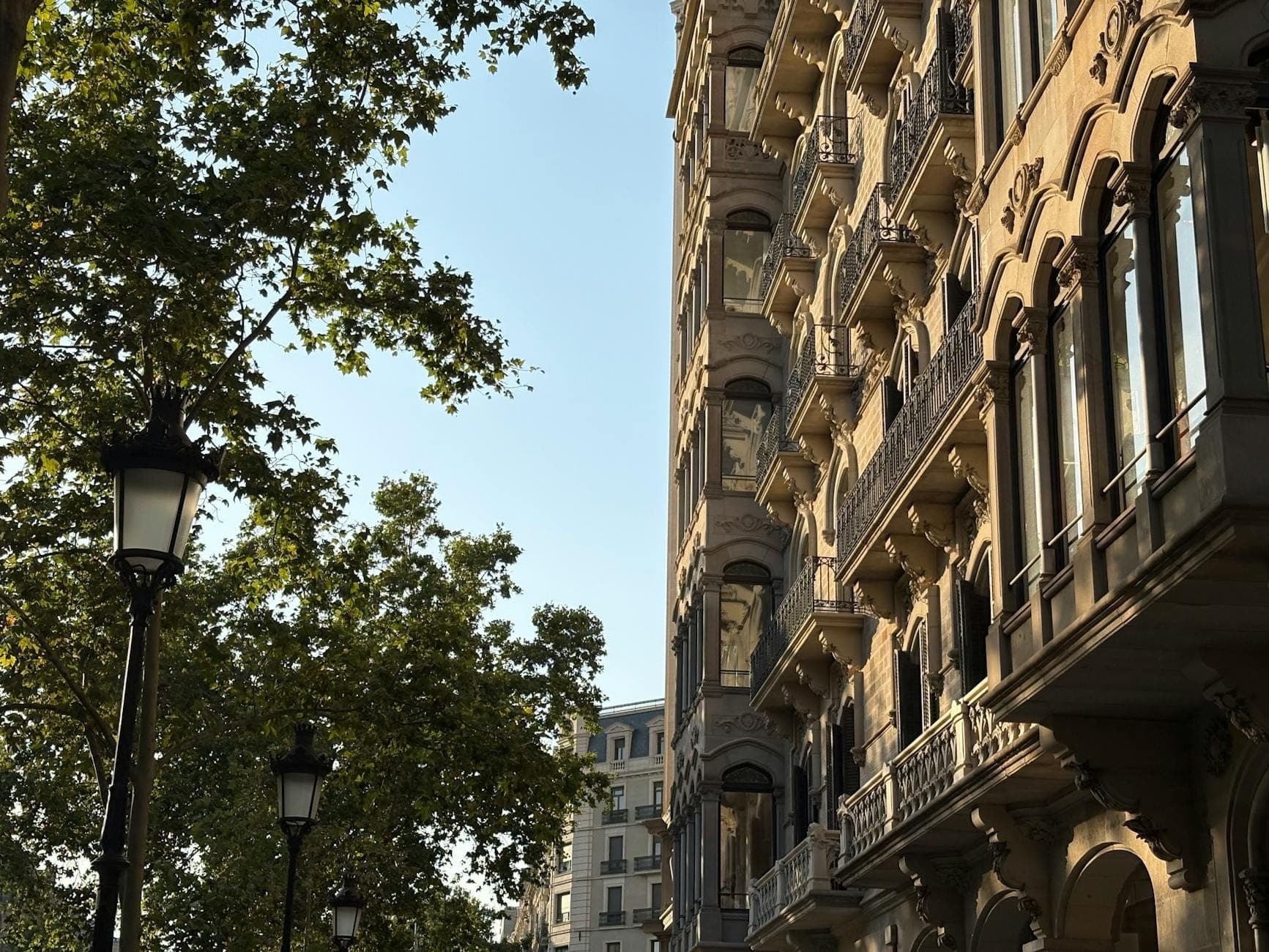 อาคารประวัติศาสตร์สง่างามและระเบียงประดับประดาเรียงรายตลอด Passeig de Gràcia ในบาร์เซโลนา พร้อมต้นไม้เขียวชอุ่มและโคมไฟถนนสไตล์วินเทจใต้ท้องฟ้าแจ่มใส