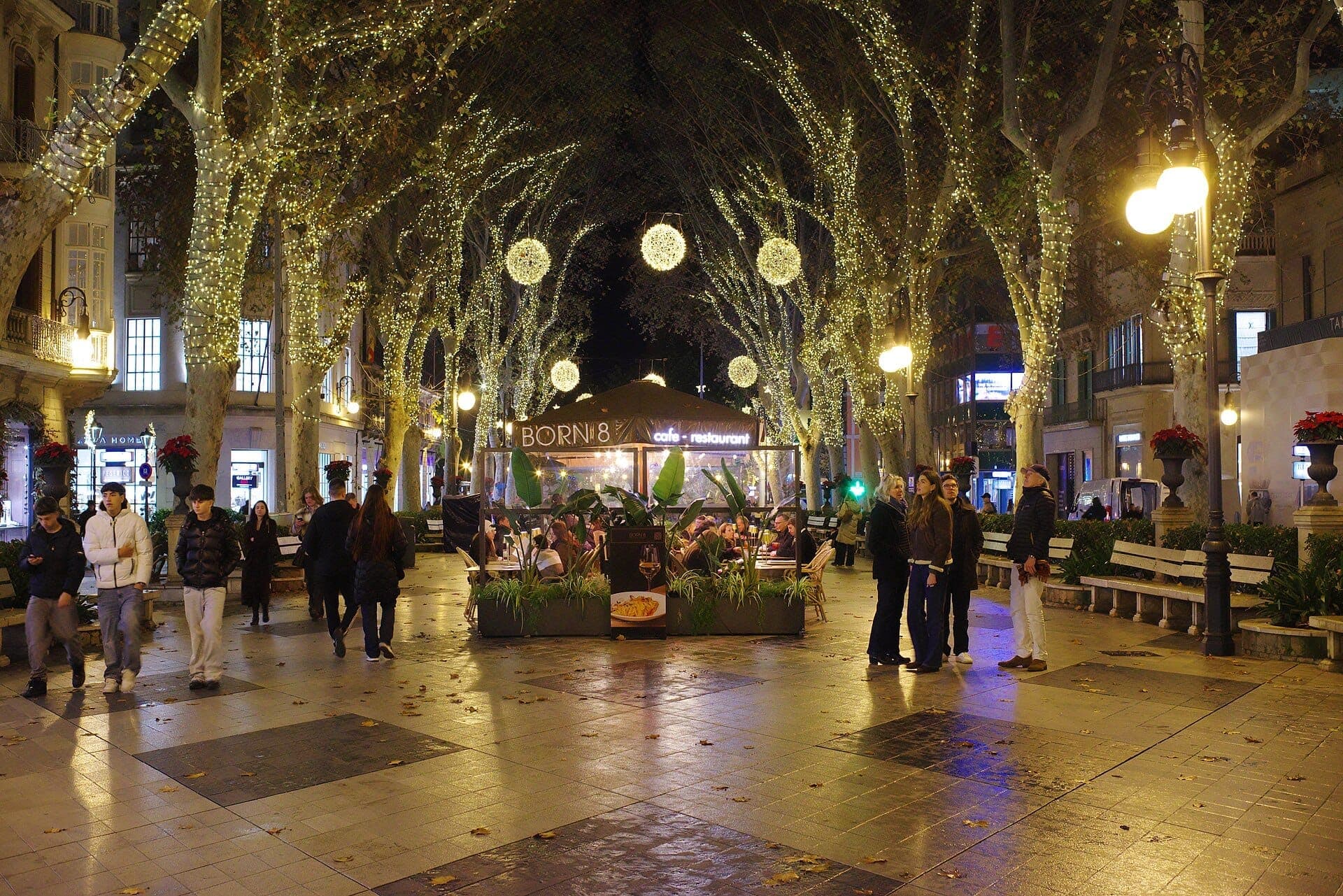 Passeig del Born bei Nacht mit beleuchteten Bäumen, belebtem Café, Menschengruppen beim Schlendern und klassischen Gebäuden entlang der Promenade.