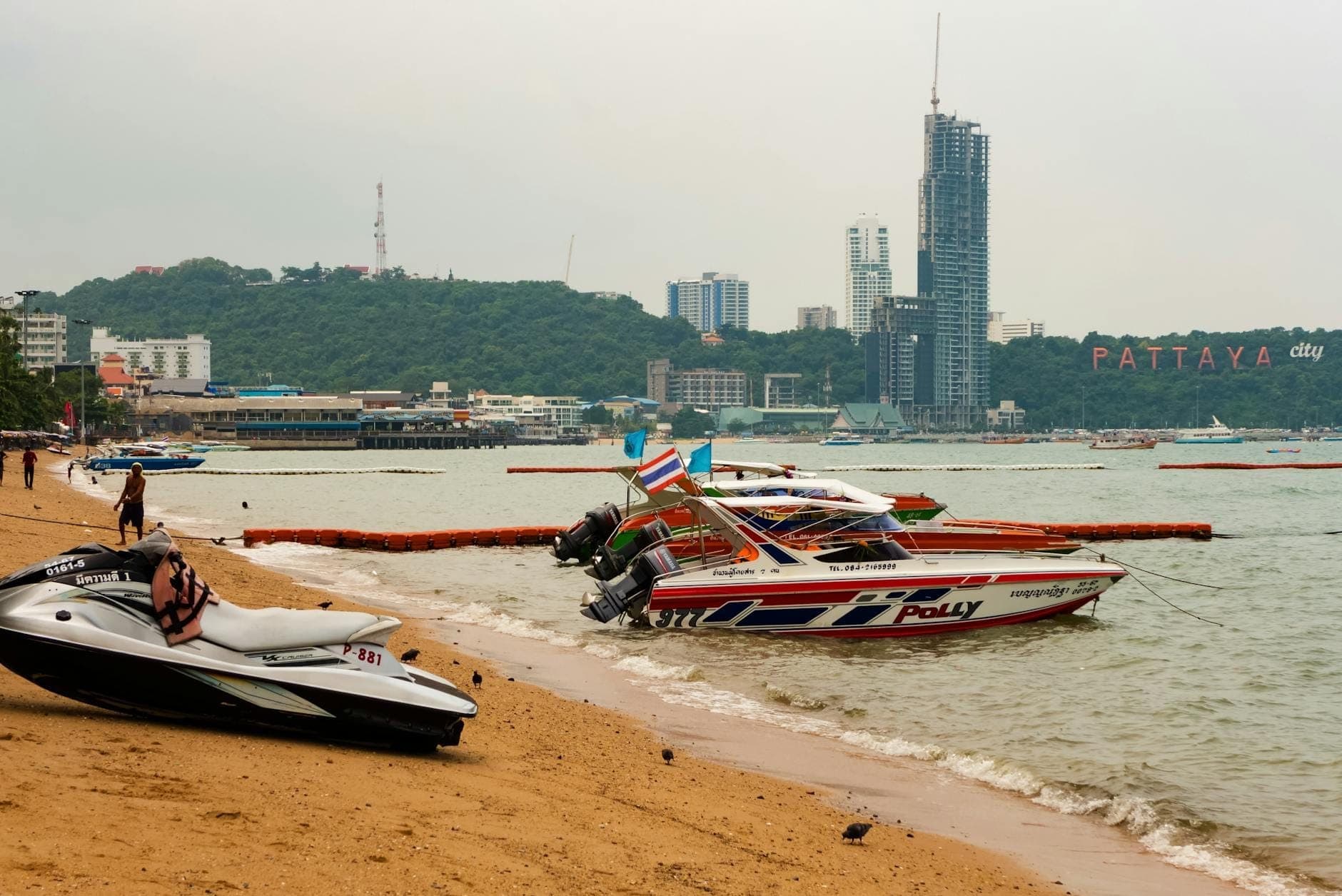 Skutery wodne i szybkie łodzie na piaszczystej plaży w Pattaya z panoramą miasta i znakiem Pattaya City w tle.