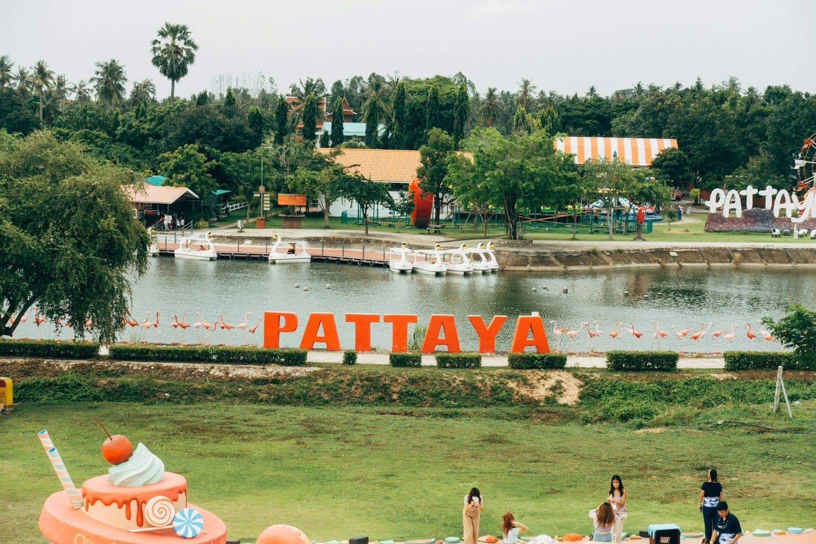 A sunny fun park in Pattaya with large 'Pattaya' letters, a lake, boats, and families and children playing on the grass in the foreground.