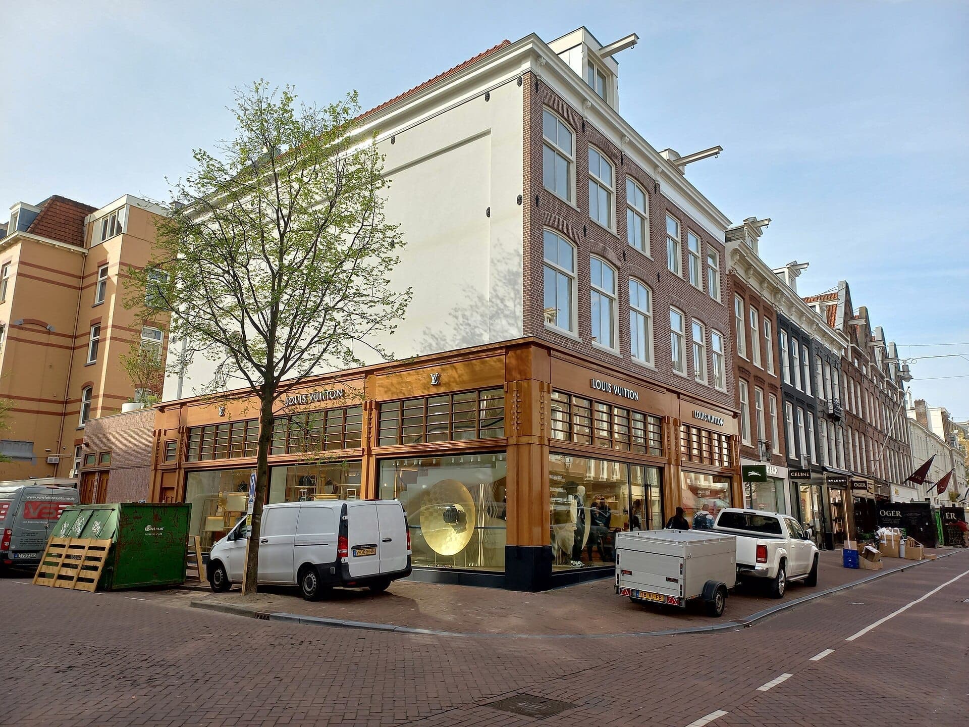Moderne Ladenfronten von Luxusmarken wie Louis Vuitton säumen die PC Hooftstraat in Amsterdam, mit großen Schaufenstern, geparkten Lieferwagen und klarem blauem Himmel.