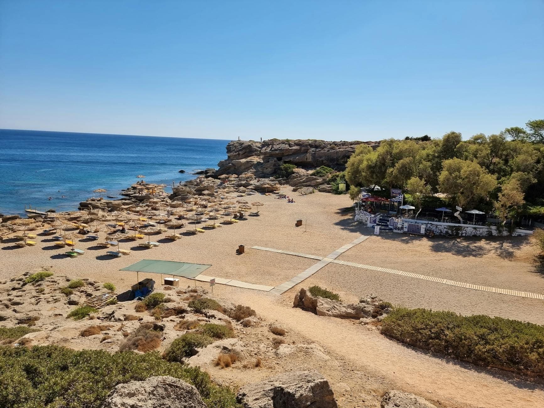 Une plage de sable tranquille avec des parasols en paille, une mer bleue et limpide, un littoral rocheux et des arbres verdoyants sous un ciel bleu éclatant, sur l'île de Rhodes.