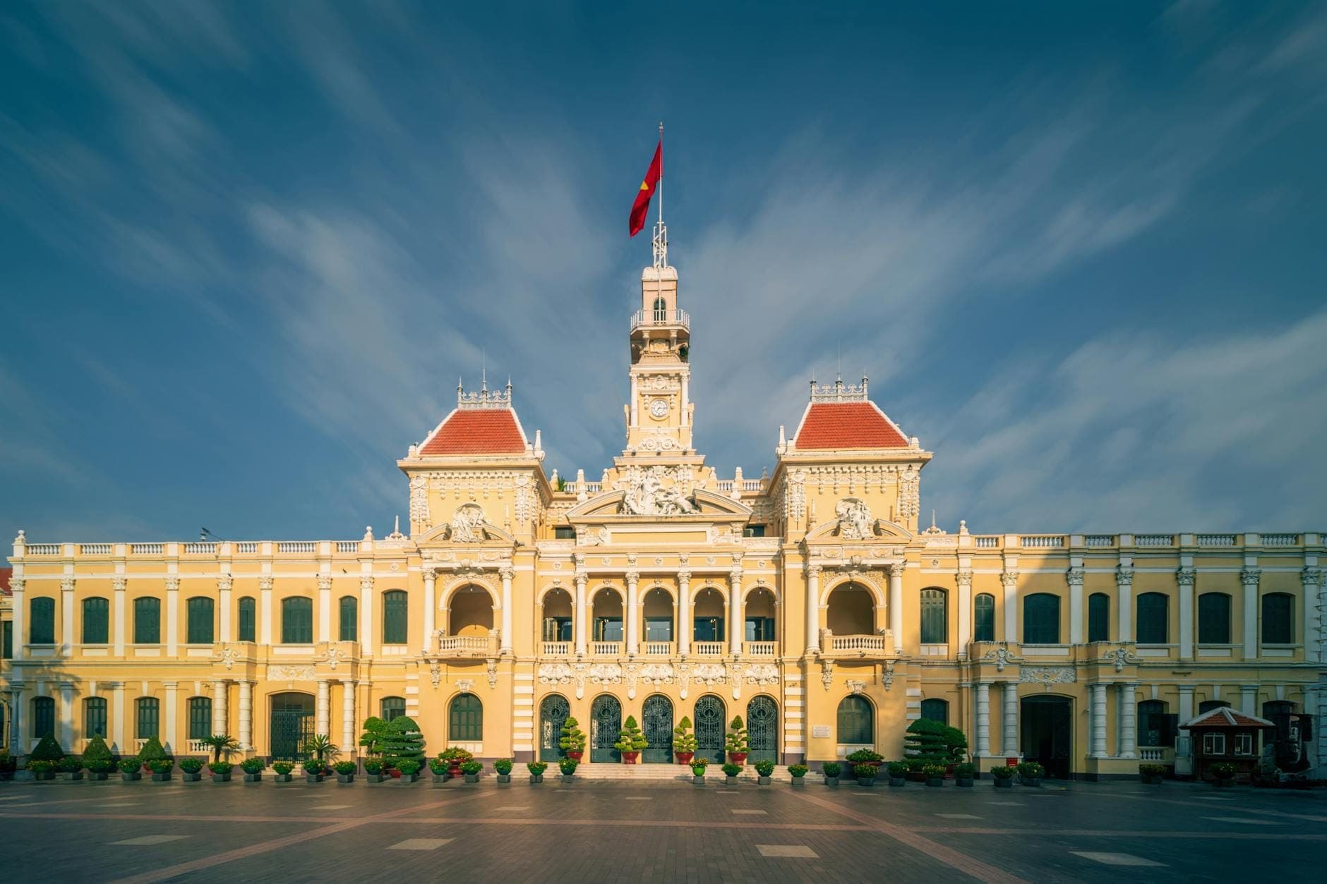 Malawak at tuwid na tingin sa People's Committee Building sa Ho Chi Minh City na may maputlang dilaw na colonial na pader, pulang bubong, at watawat ng Vietnam sa ilalim ng dramatikong asul na langit.