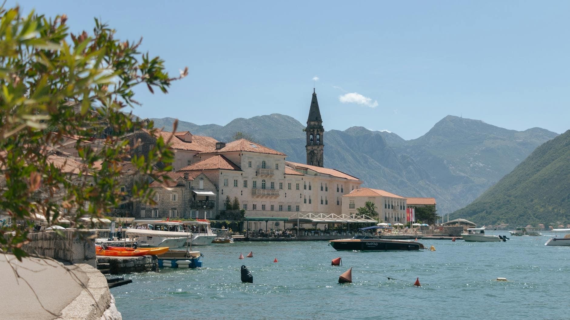 Vue ensoleillée de la vieille ville de Perast avec ses bâtiments baroques, ses cafés au bord de l'eau, des bateaux sur la baie de Kotor et les montagnes en arrière-plan.