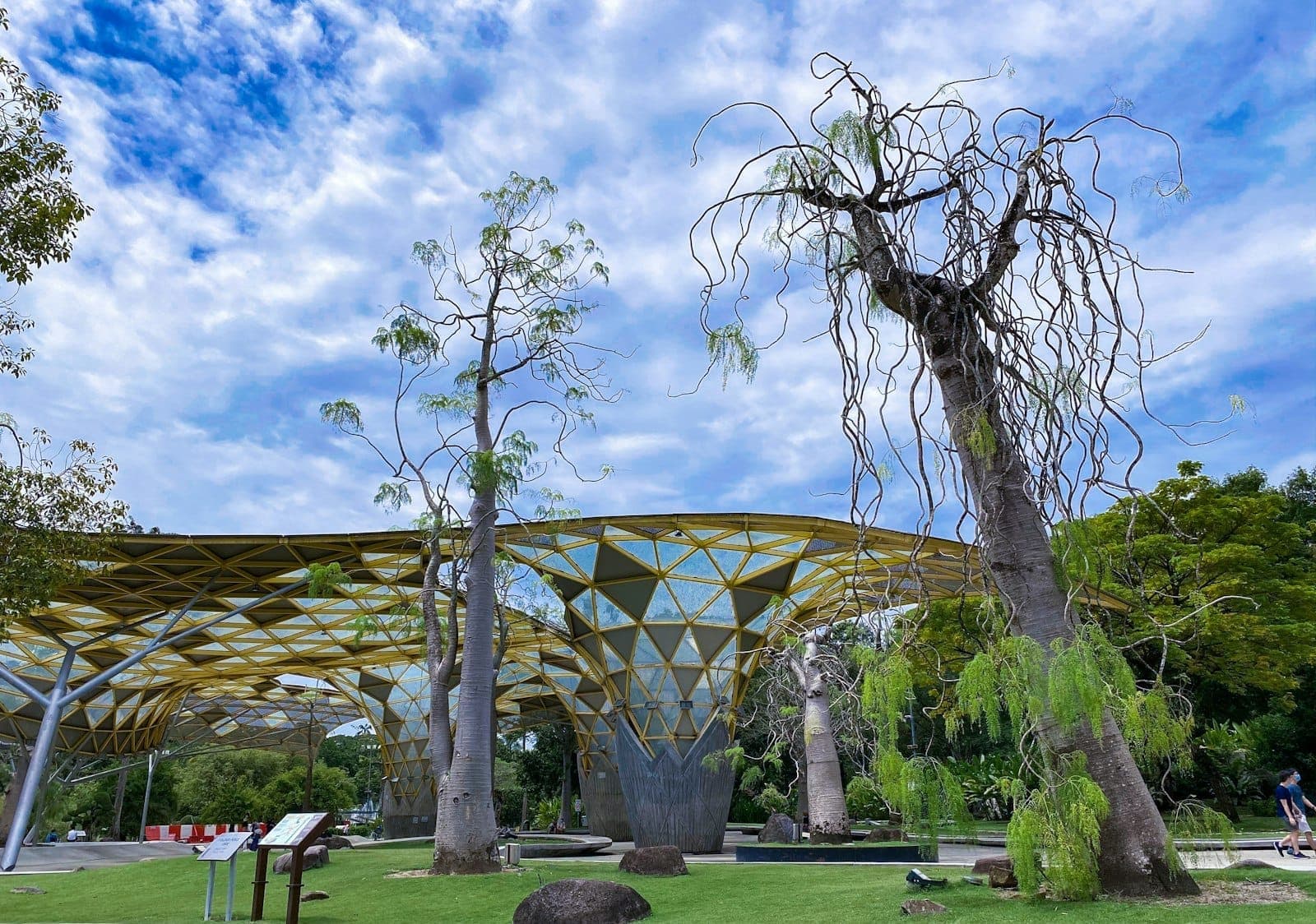 Perdana Botanical Gardens, Kuala Lumpur'da geometrik sarı tente yapıları ve benzersiz ağaçlar, parlak mavi gökyüzü ve dağınık bulutlar altında.