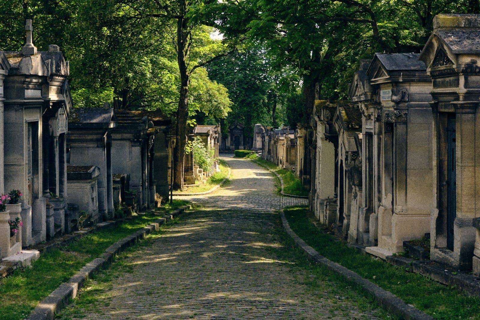 Sunlit cobblestone path winding between ornate tombs and mausoleums beneath leafy green trees at Père Lachaise Cemetery in Paris, evoking peace and history.