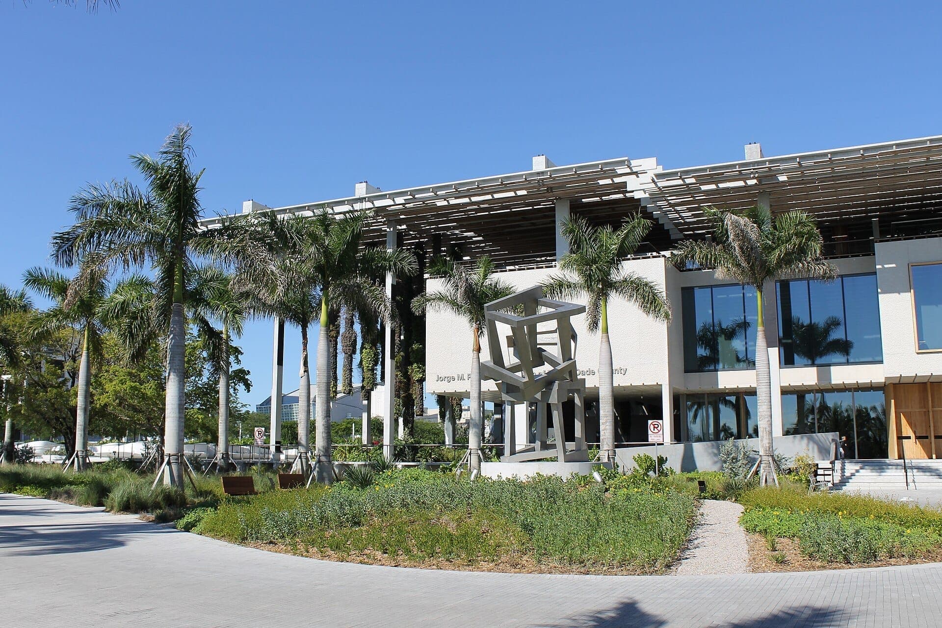 Front view of Pérez Art Museum Miami with palm trees, modern architecture, glass windows, and a geometric metal sculpture under a clear blue sky.