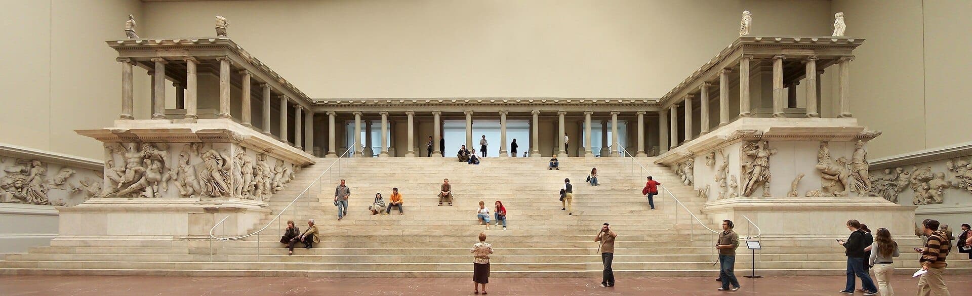Монументальный Пергамский алтарь внутри Pergamon Museum в Берлине: посетители осматривают величественные мраморные ступени и искусные античные фризы в мягком внутреннем освещении.