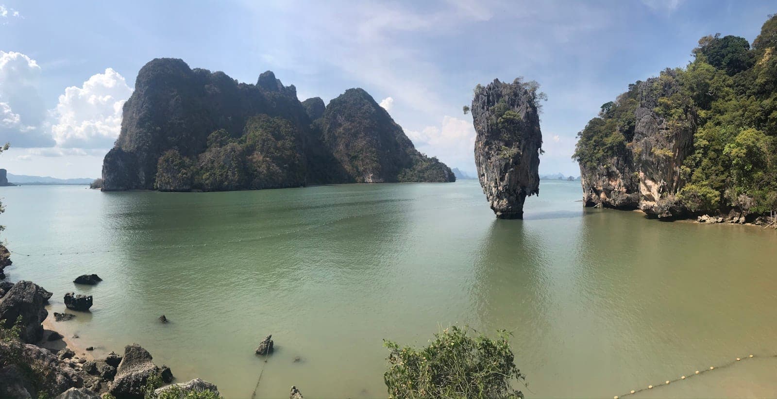 Wide panoramic view of Phang Nga Bay with the iconic James Bond Island limestone karst rising from calm green waters and lush cliffs under a bright sky.