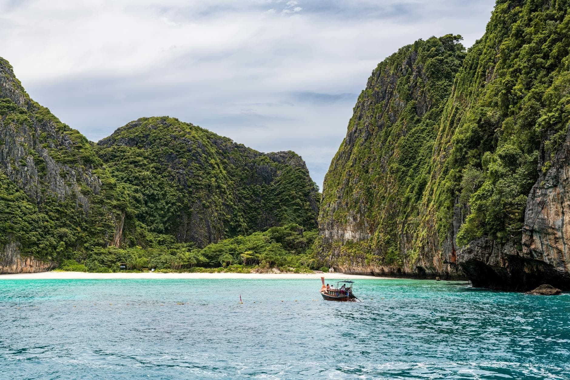Turquoise water and a traditional longtail boat approaching a white sand beach surrounded by dramatic limestone cliffs at the Phi Phi Islands.