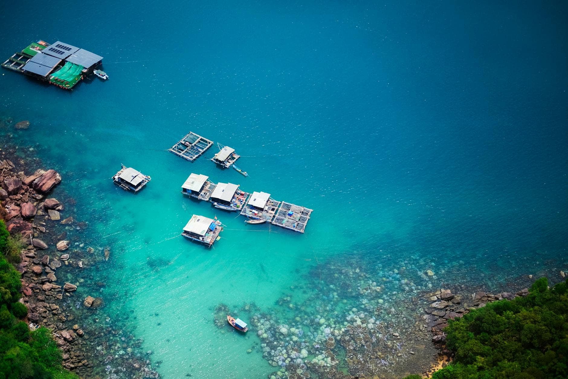 Cảnh nhìn từ trên cao của vùng nước xanh ngọc với các cầu tàu nổi và thuyền nhỏ gần bờ biển đá cây xanh, gợi lên khung cảnh hòn đảo thiên nhiên yên bình.